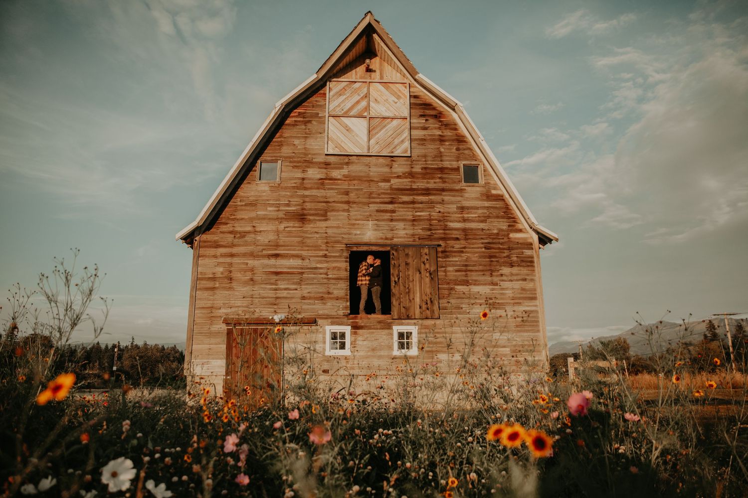 A rustic wooden barn surrounded by wildflowers and sunflowers under a moody sky at sunset.