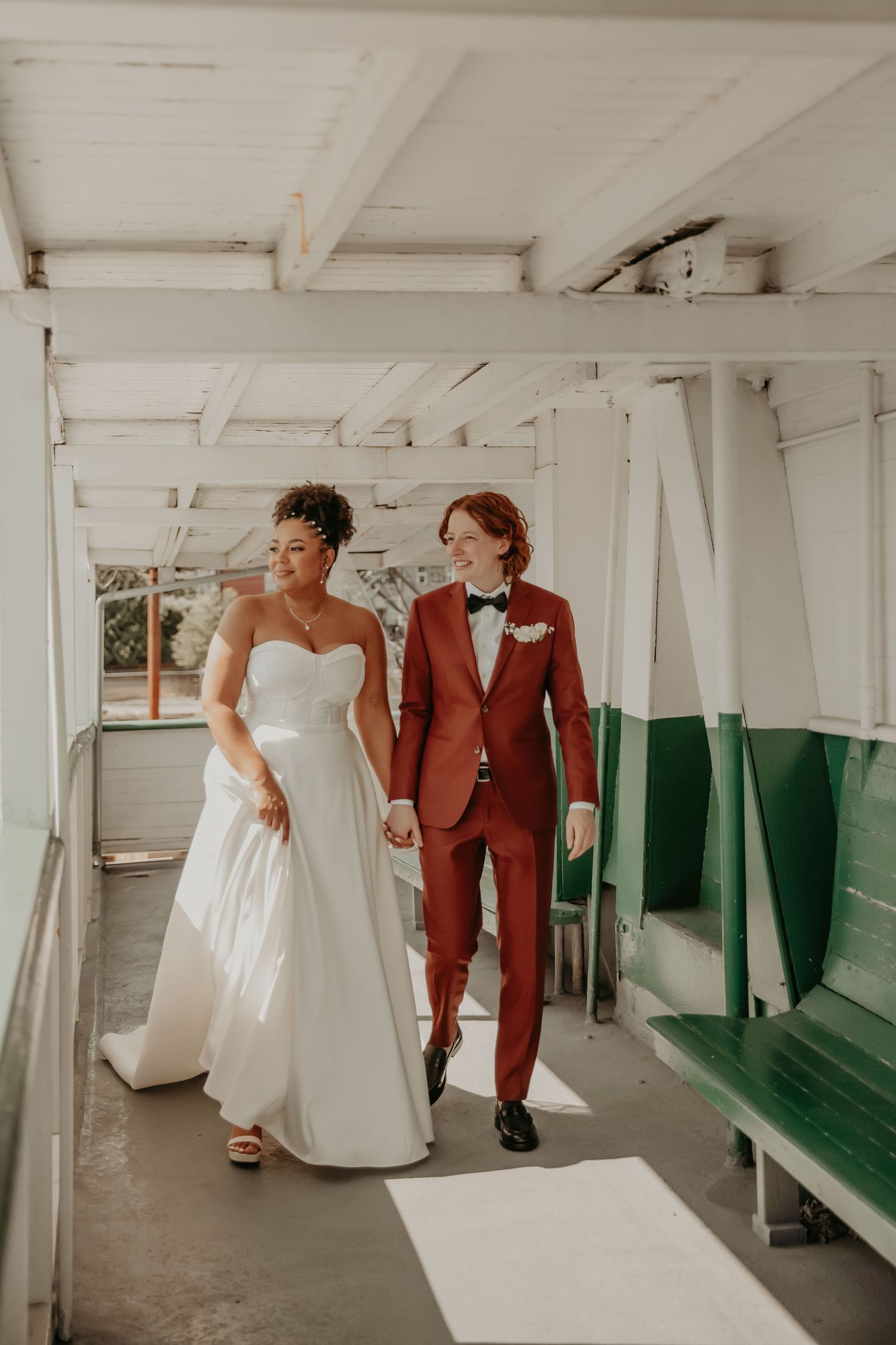 Two newlyweds walking together on a boat, one in a white wedding gown and one in a rust-colored suit.