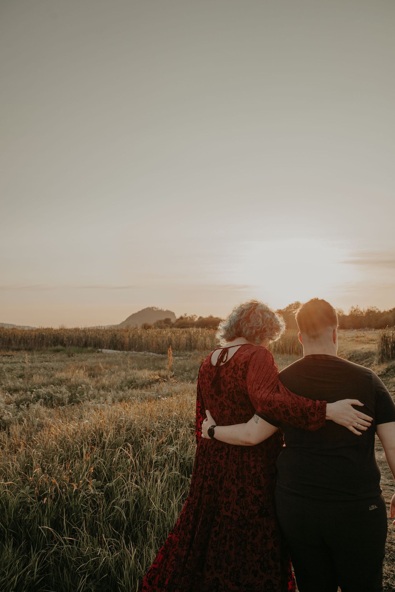 Couple embracing in a field at sunset with mountains in background and person wearing red flowing dress.