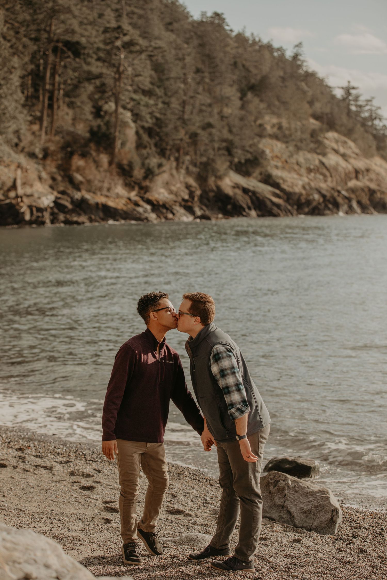 Couple sharing an intimate moment on a rocky beach with tall cliffside and evergreen trees in the background.