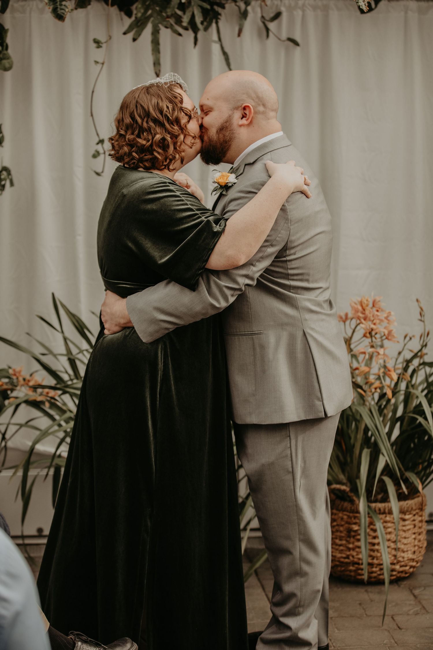A romantic moment captured as a couple in formal attire share a kiss against a backdrop of greenery.