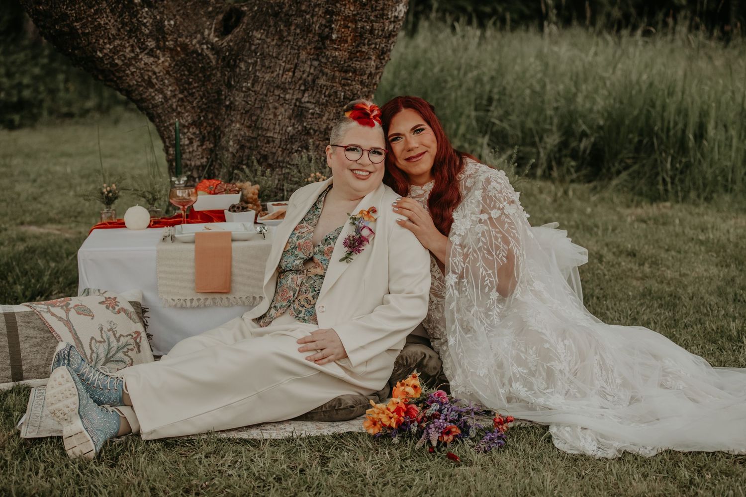 A romantic outdoor picnic wedding scene with couple sitting under a tree surrounded by floral arrangements and decor.