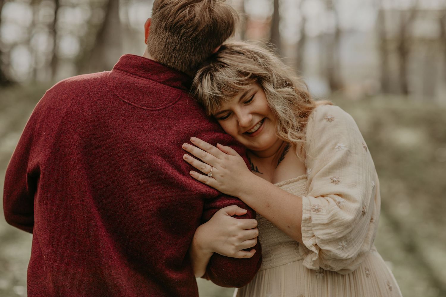 A romantic couple embraces in an autumn forest setting, with warm burgundy and cream colored clothing.