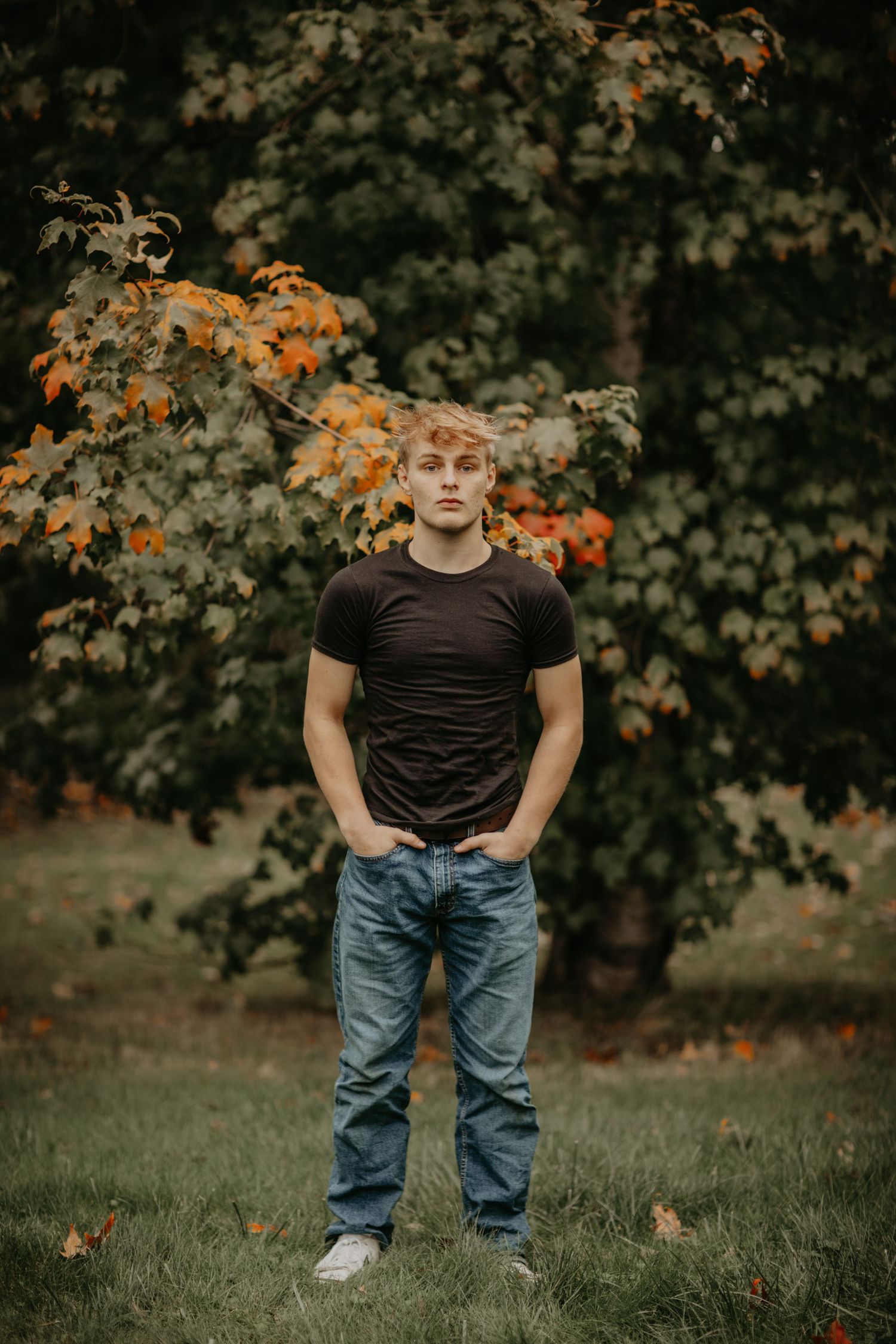 A person stands casually with hands in pockets against autumn foliage and a blurred tree background.