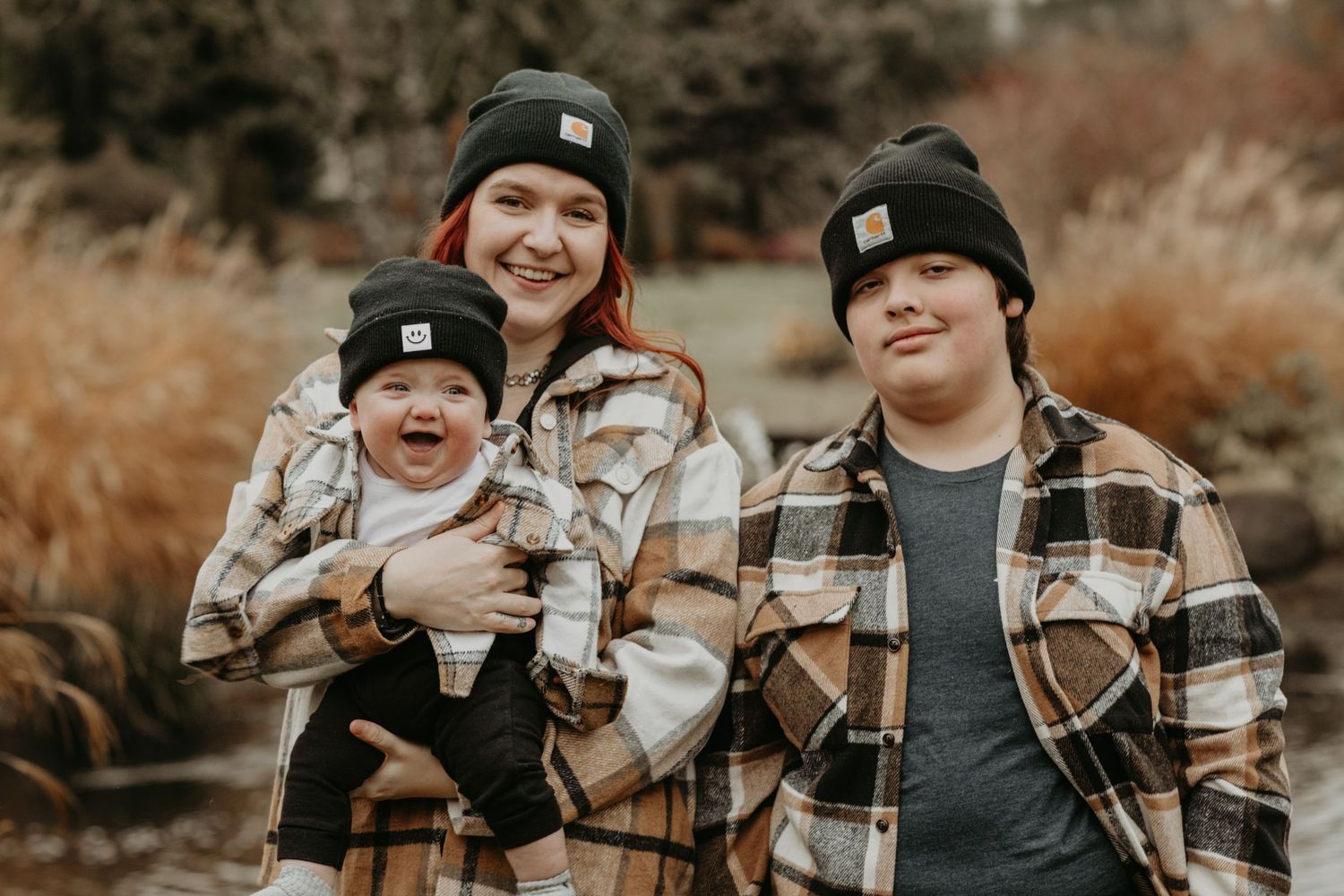 Three people wearing winter beanies and plaid flannel clothing pose together outdoors during autumn.