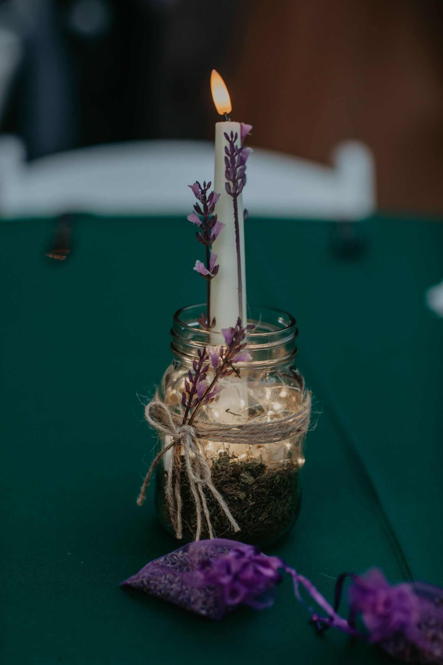 A lit white candle decorated with purple lavender sprigs in a rustic mason jar centerpiece on a green tablecloth.