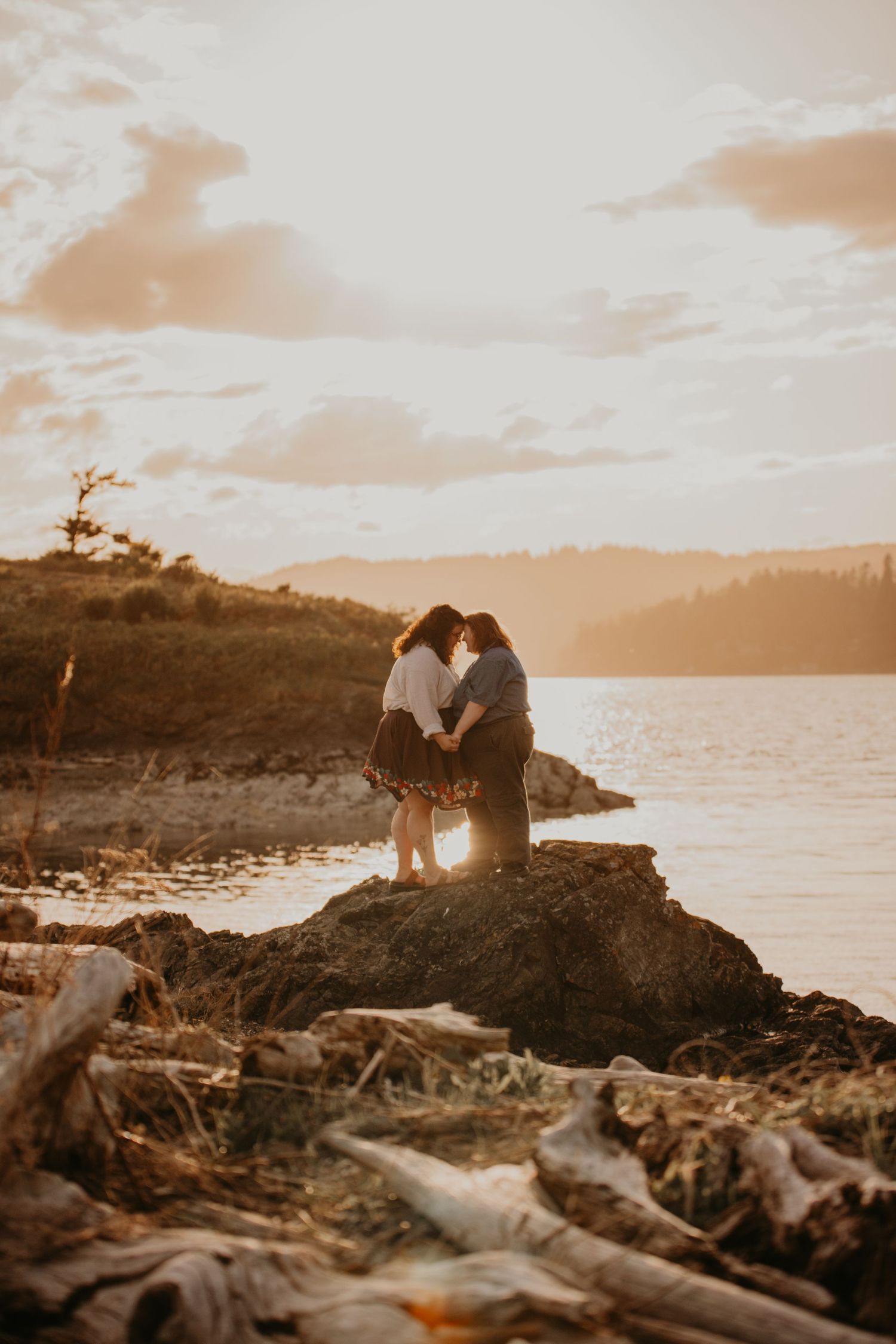A romantic sunset silhouette of a couple standing together on rocky coastal shore with warm golden light over the water.