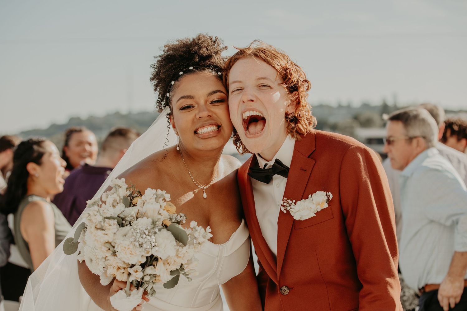 A joyful newlywed couple laughs together outdoors, with one wearing a rust-colored suit and the other in a white gown.