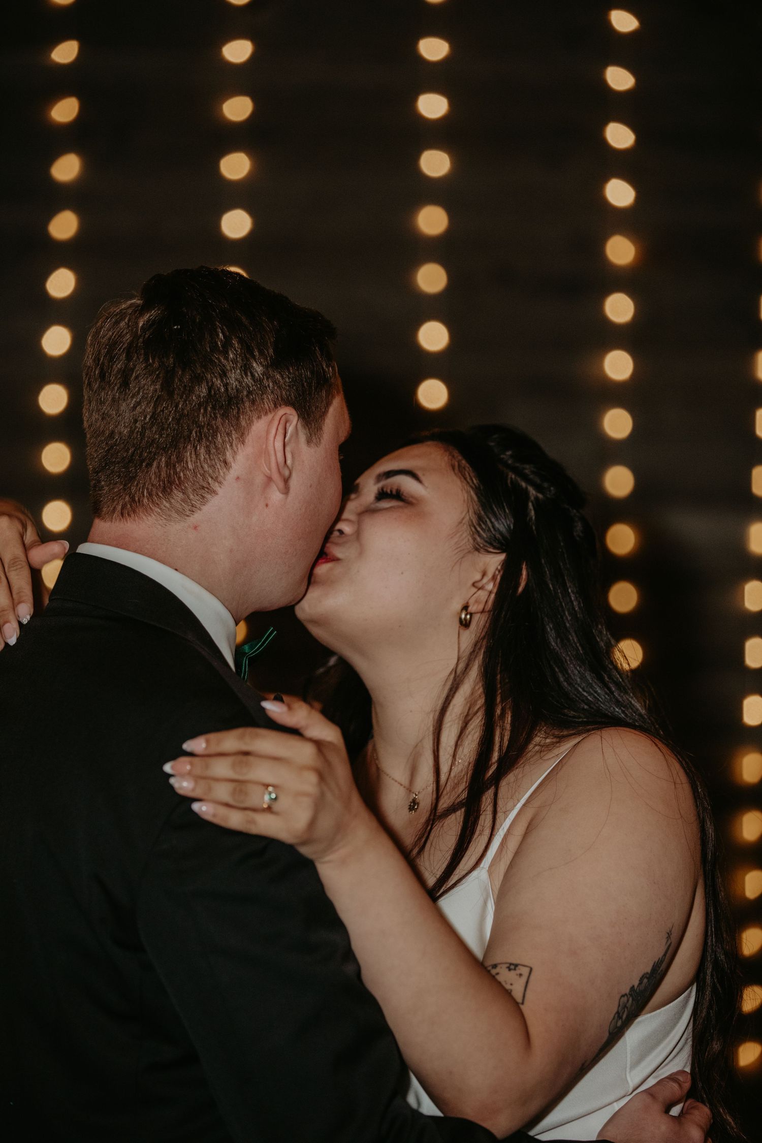 A romantic kiss between newlyweds against a backdrop of warm glowing string lights at an evening wedding celebration.