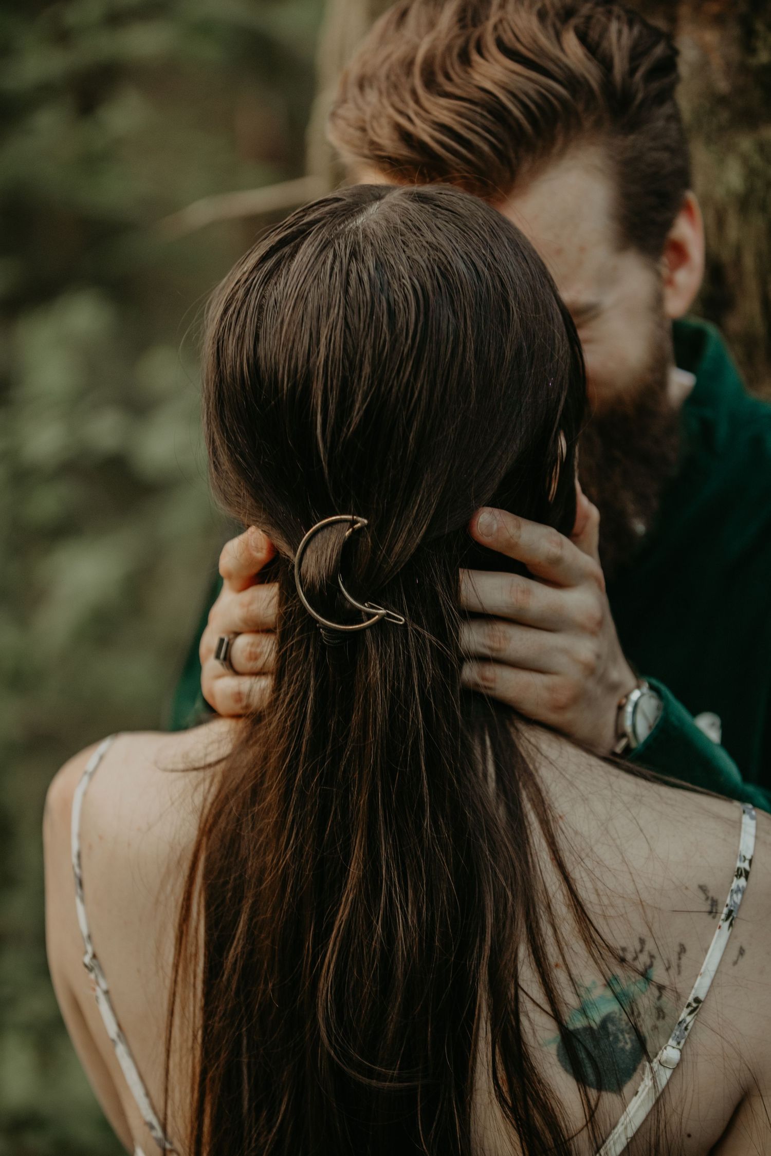 Intimate outdoor portrait showing long dark hair with crescent moon hairpin and delicate dress straps against blurred green backdrop.
