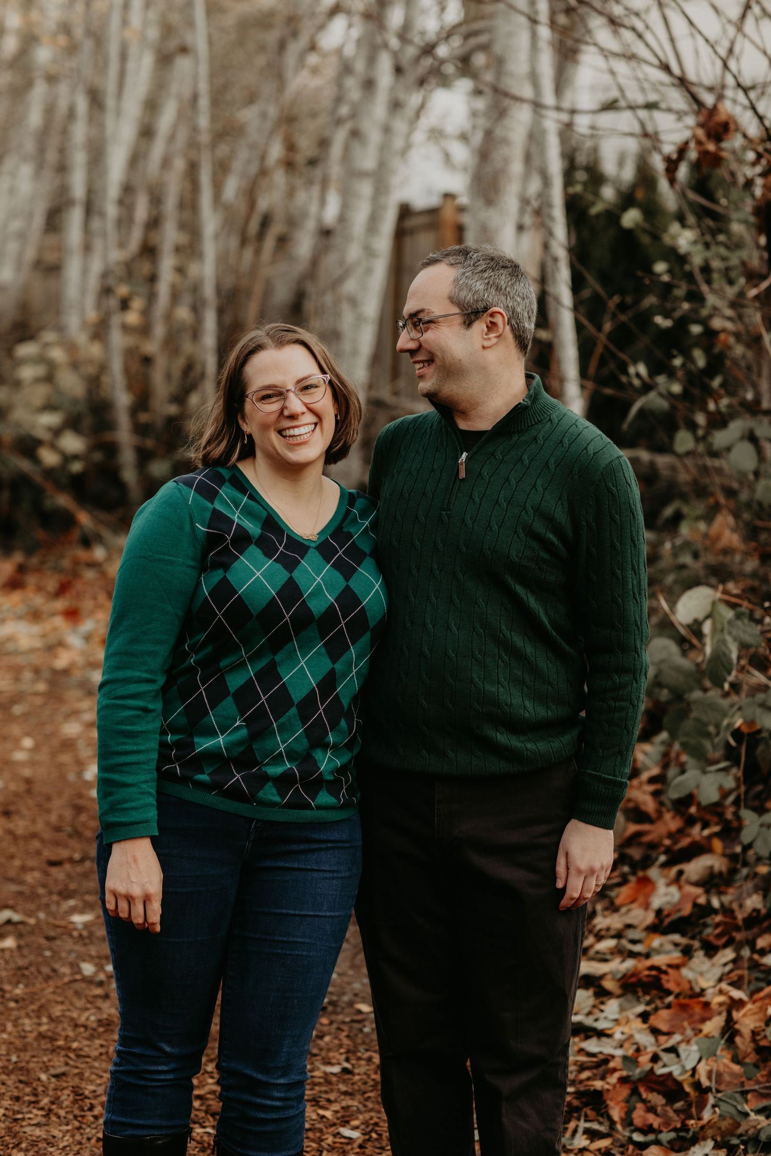 A couple wearing matching green sweaters shares a laugh together in a wooded autumn setting.