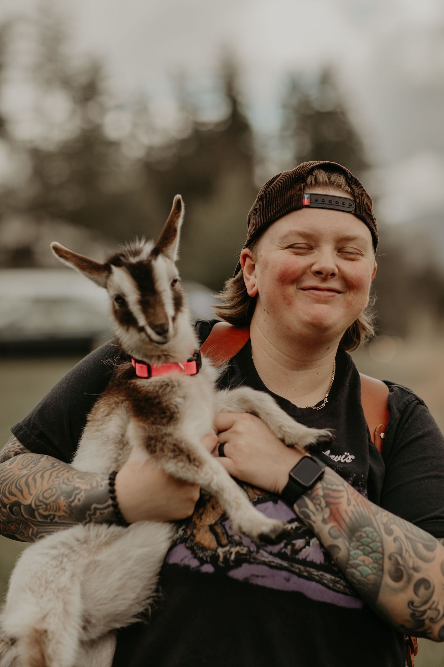 An adorable baby goat with black and white markings is being held by someone wearing a baseball cap and tattoos.