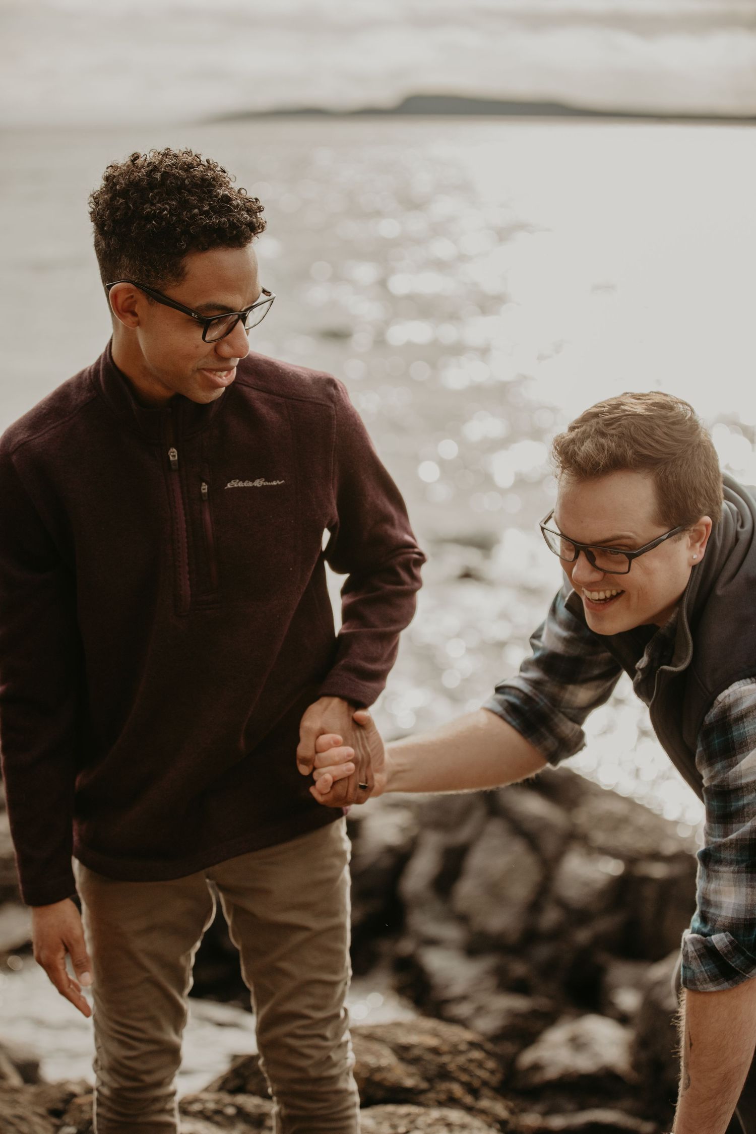 Two friends wearing glasses and casual clothing share a lighthearted moment while standing on rocky terrain near water.