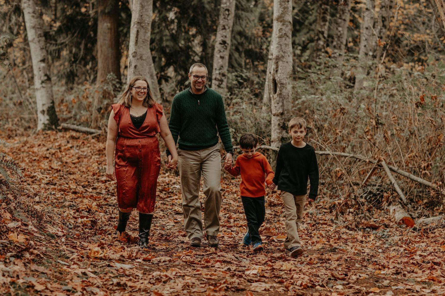 A family walks together through a forest path covered in fallen autumn leaves while wearing cozy fall attire.