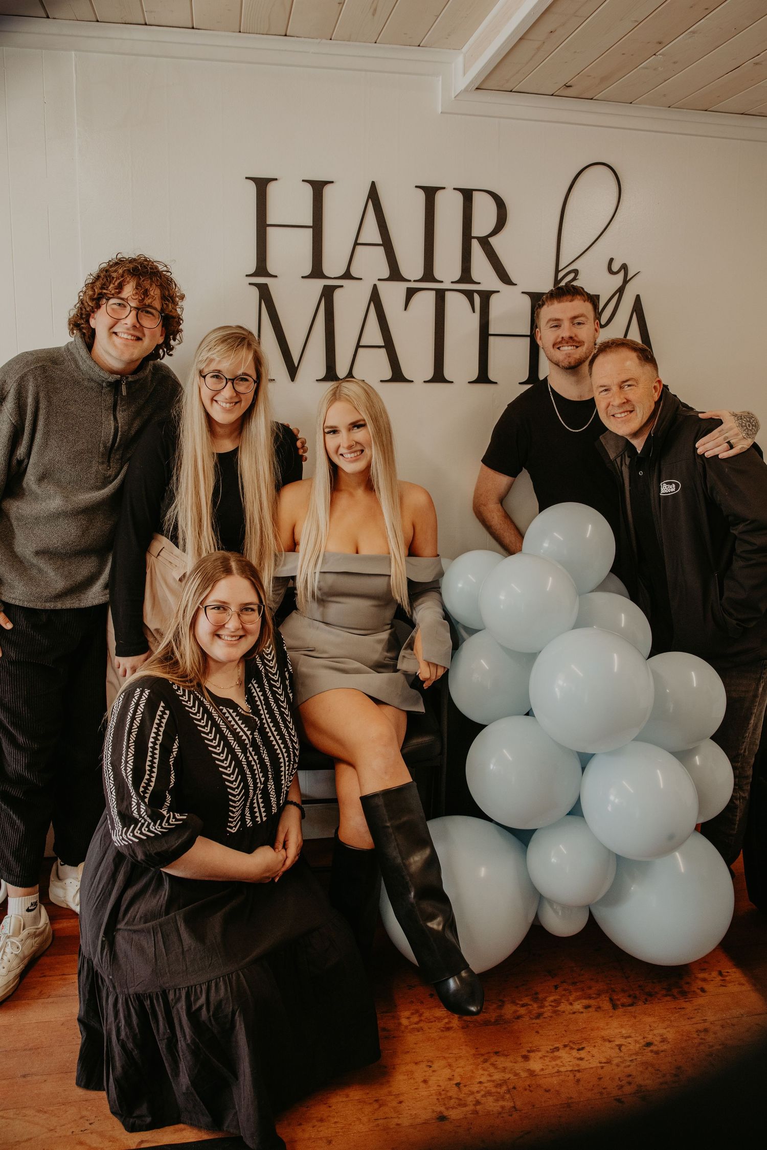 A group of salon team members pose together by a 'Hair by Math' wall sign with light blue balloon decorations.