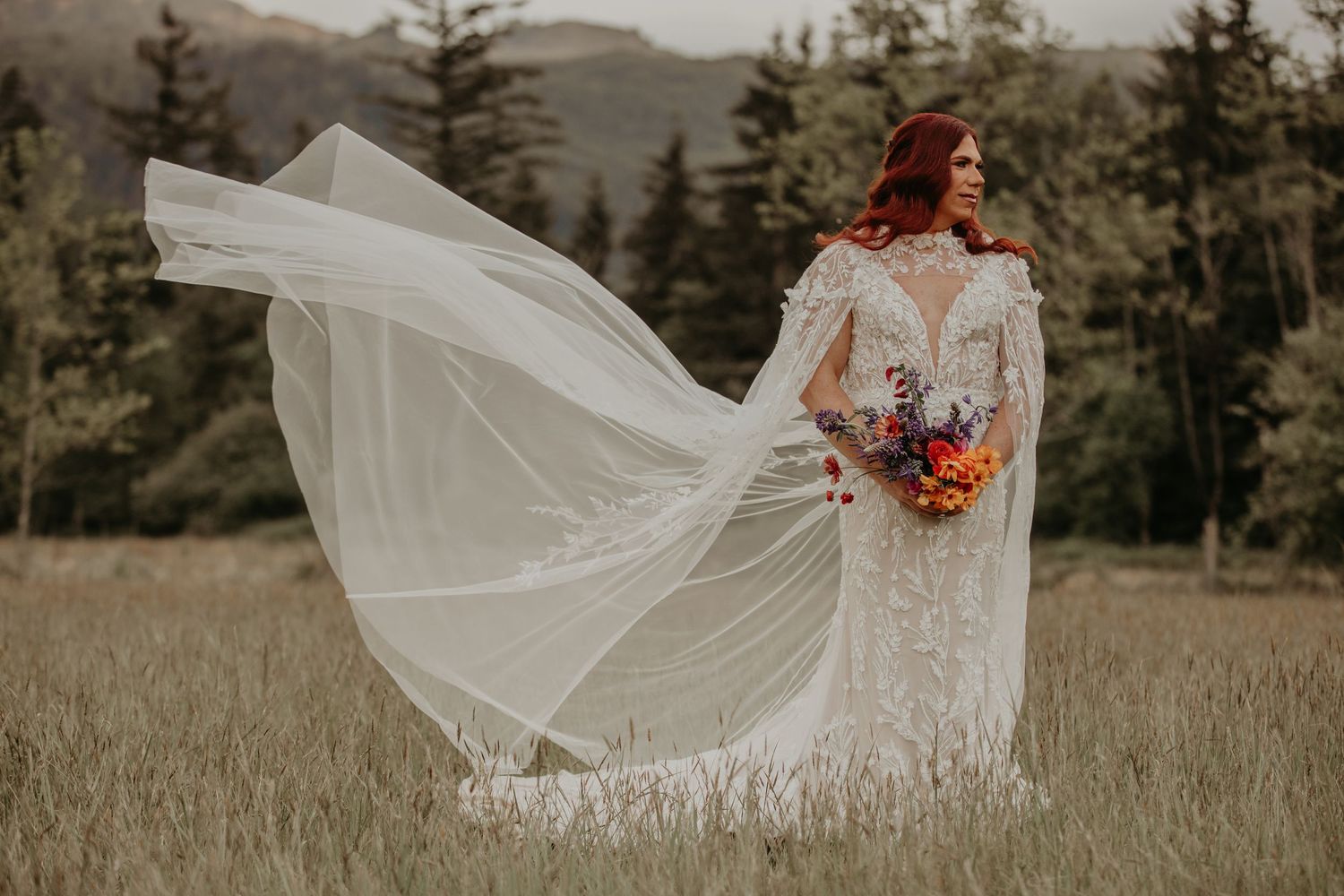 A bride in a lace dress holds a colorful bouquet while her veil dramatically flows in the wind against a mountain landscape.