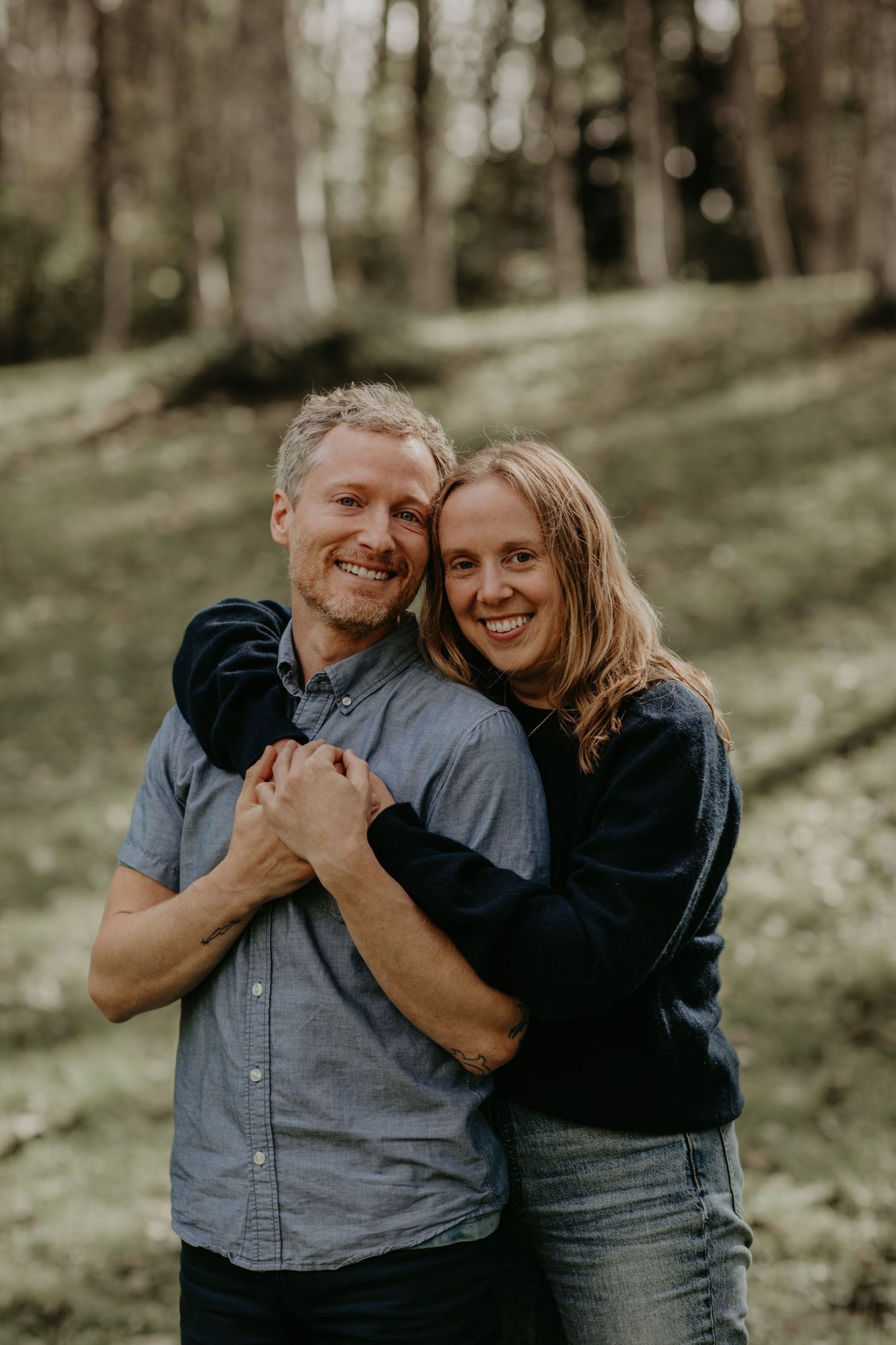 A couple embraces while standing on a wooded hillside in warm natural lighting during an outdoor photo session.