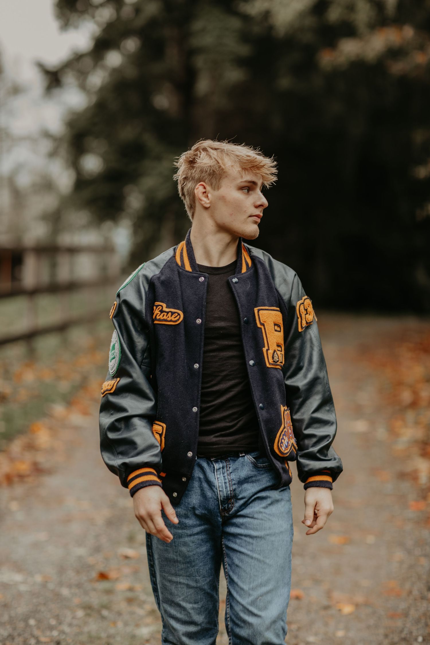 Person wearing a dark varsity letterman jacket with orange patches and blue jeans poses on an autumn path.