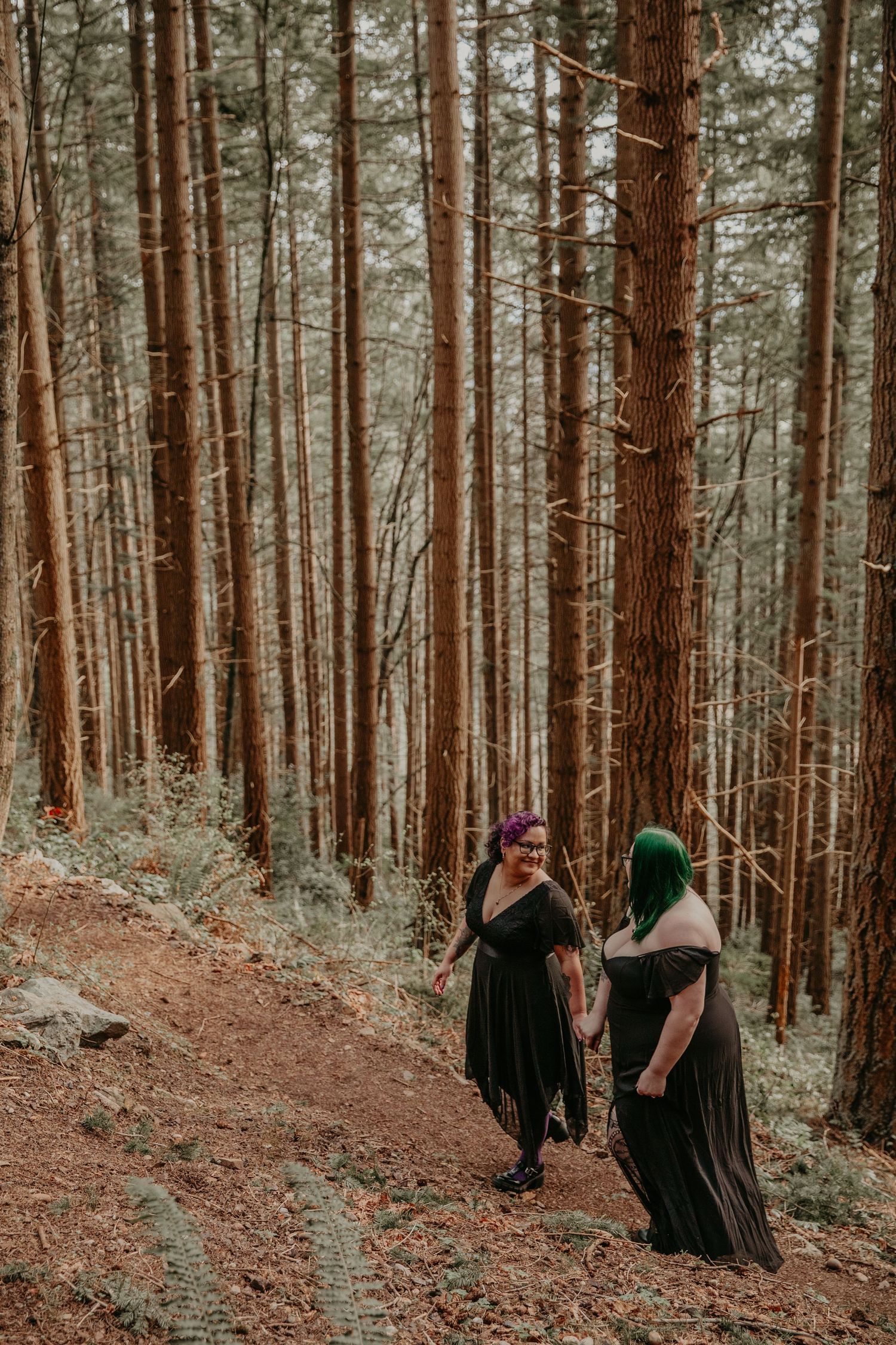 Two people in long black dresses walking together on a forest trail surrounded by tall redwood trees in warm lighting.