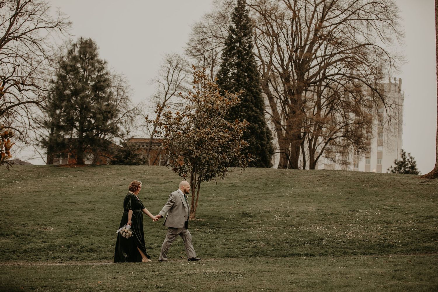 Two figures walk up a grassy hillside together on an overcast day surrounded by bare trees in the background.