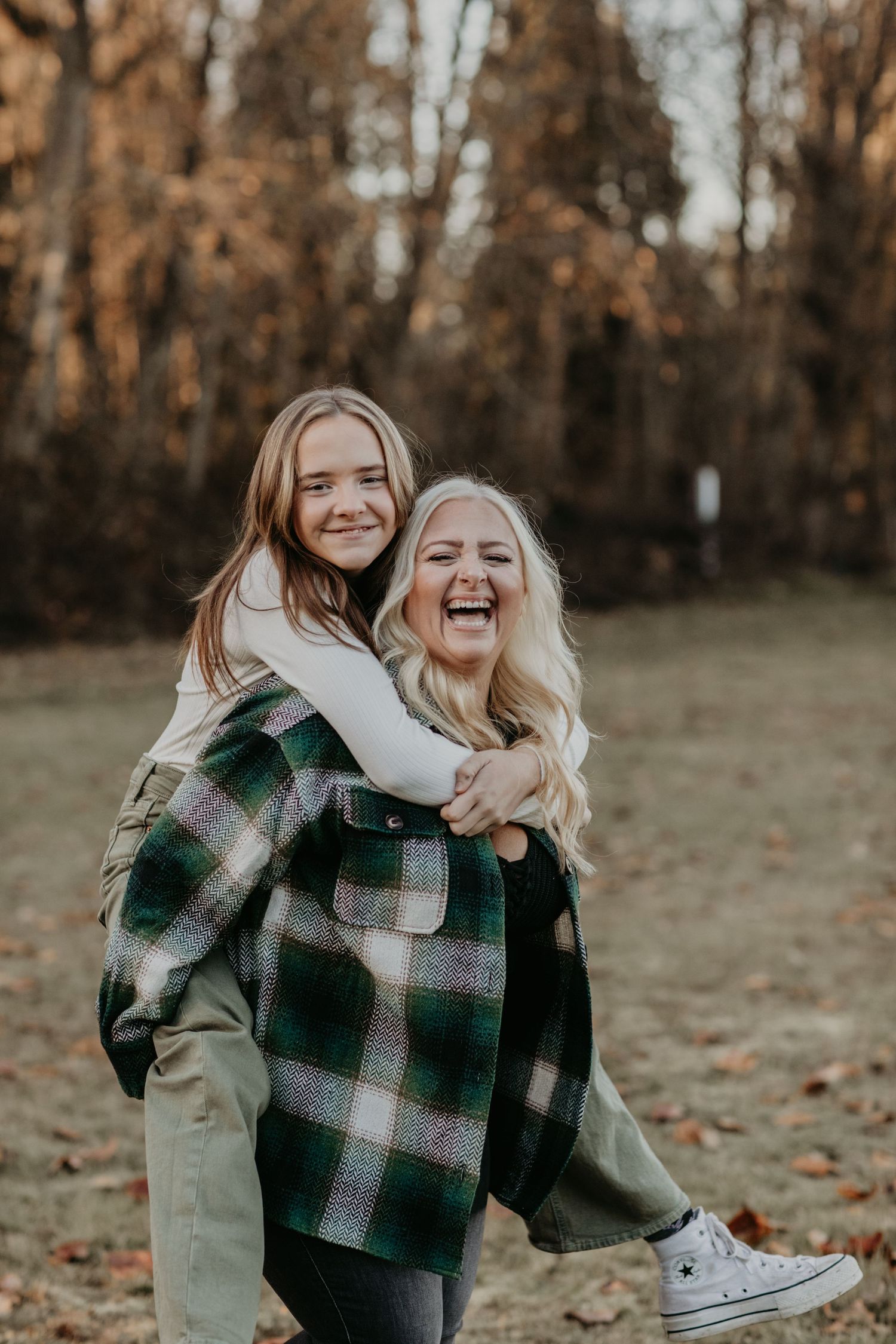Friends laugh together during a playful piggyback ride in an autumn forest setting with fallen leaves.