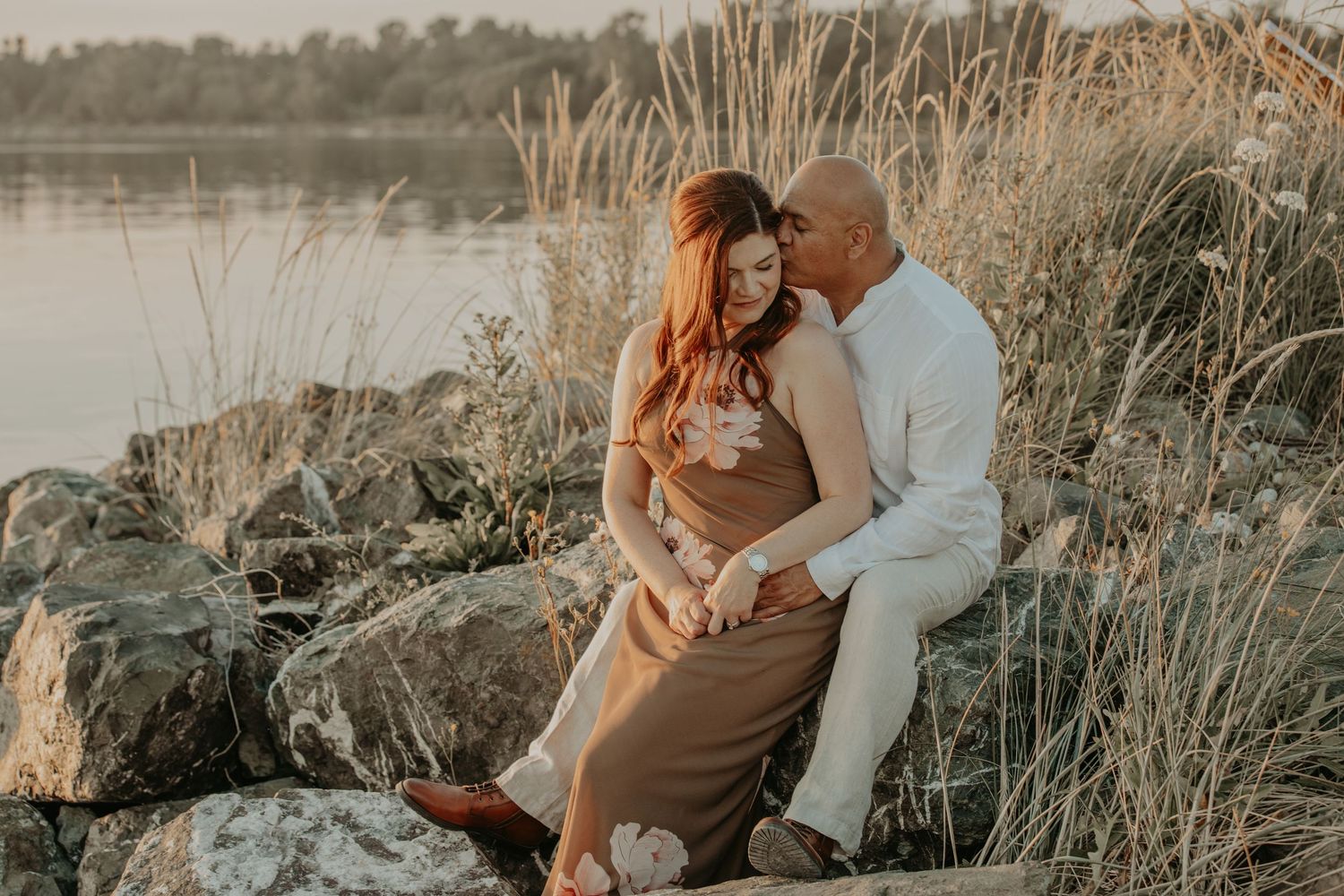 A couple embraces during golden hour by a scenic lake with tall grasses and warm sunset lighting in the background.