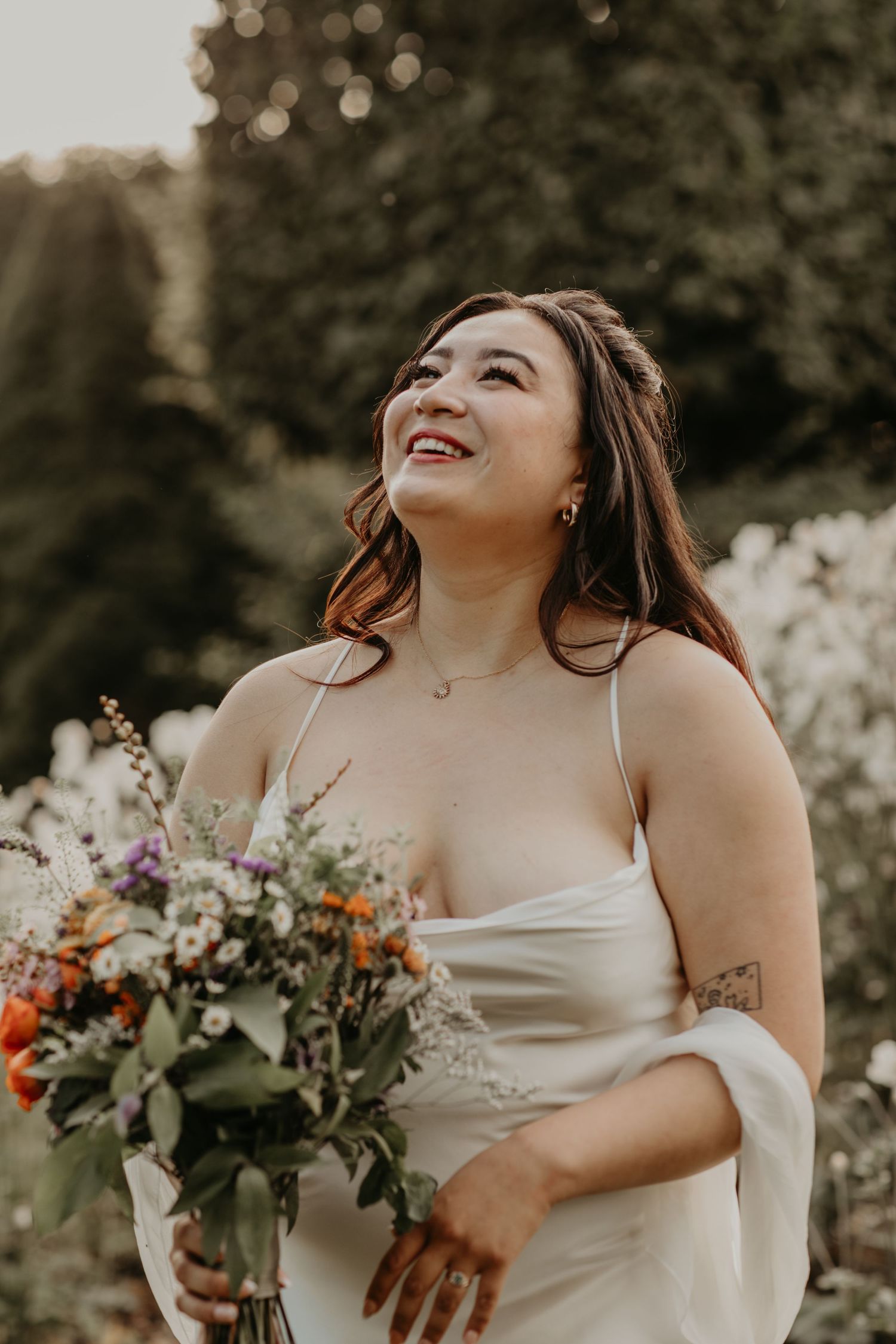 A person in a white wedding dress holds a vibrant wildflower bouquet while smiling joyfully in golden natural light.