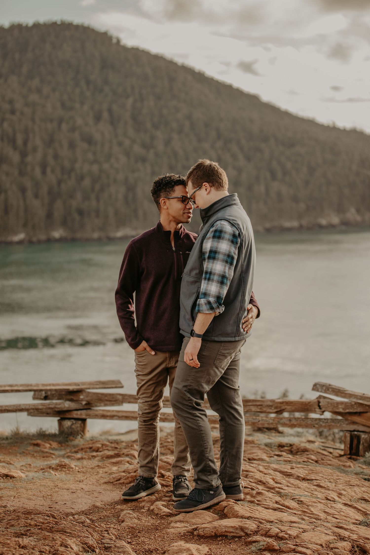 A couple shares an intimate moment on a cliff overlooking a serene lake surrounded by forested mountains at sunset.