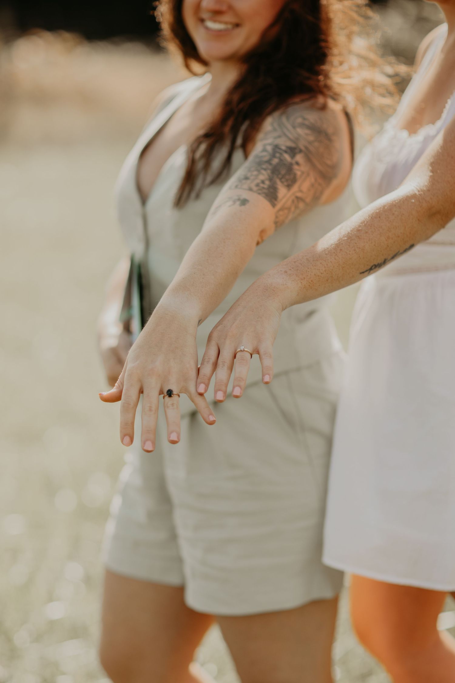 Two hands showing off engagement rings with light streaming in and one person wearing a sleeveless romper with visible tattoo.