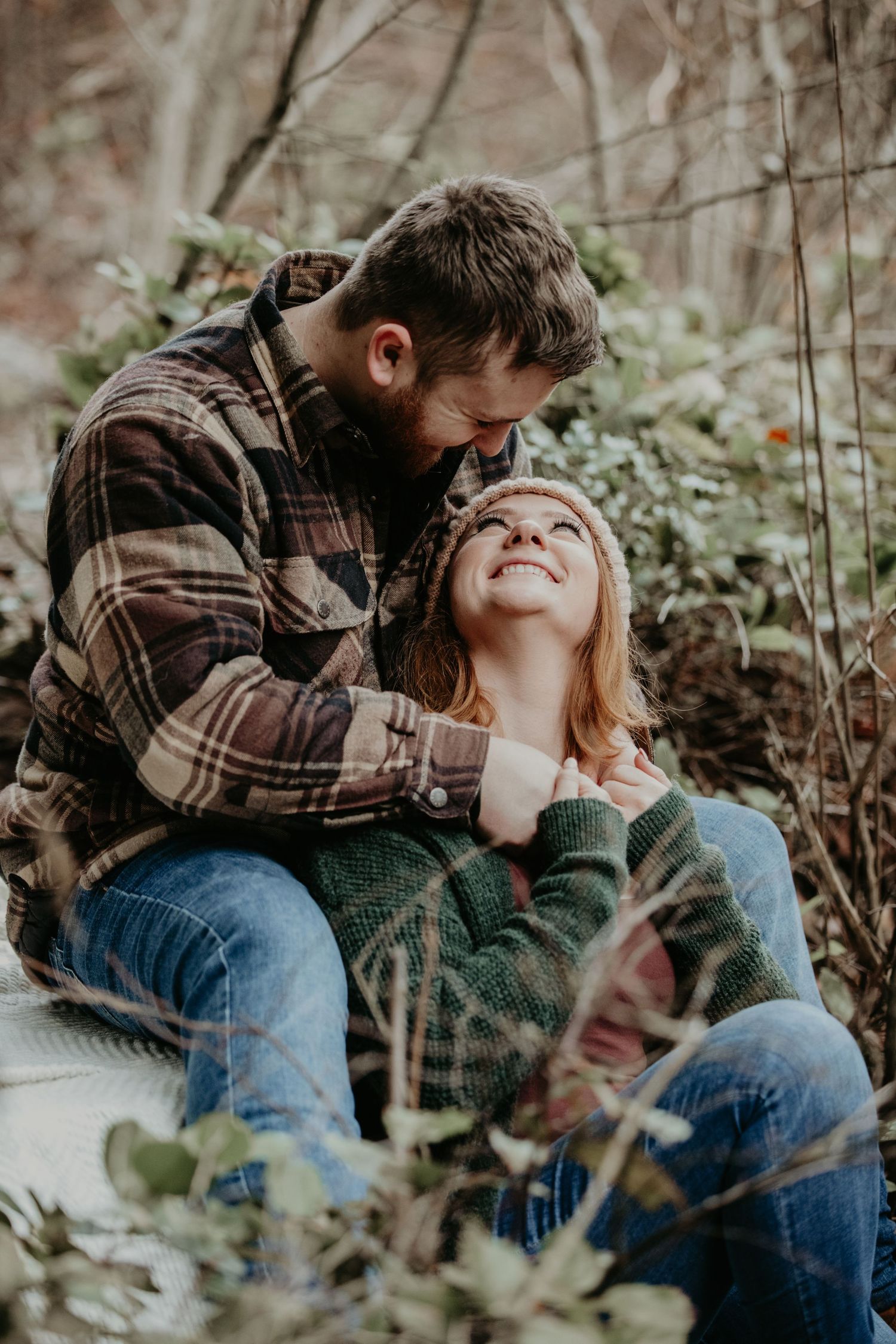 A couple in casual clothing sits together outdoors among foliage in an intimate autumn moment.