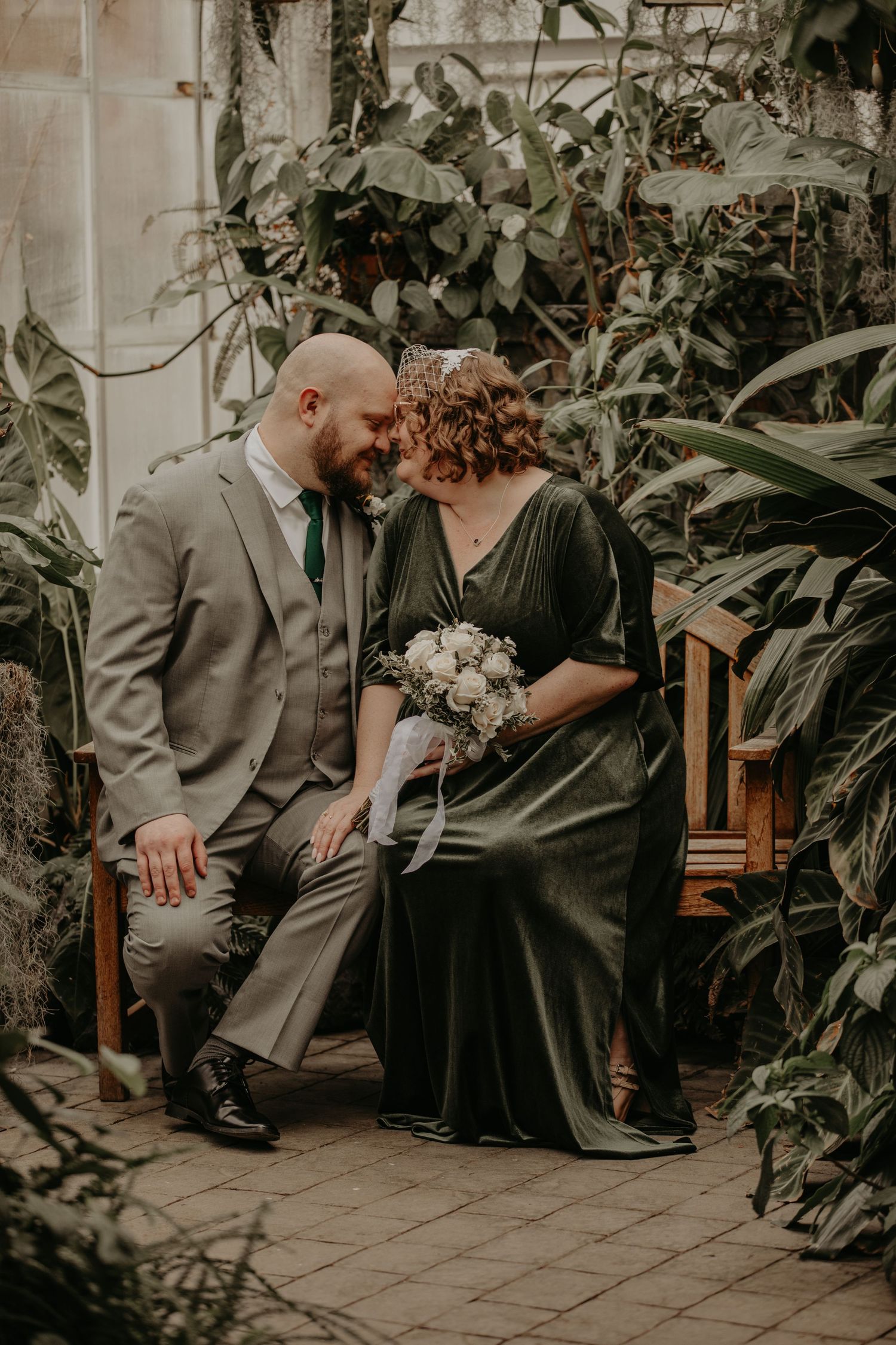 A couple in wedding attire share an intimate moment on a wooden bench surrounded by lush greenhouse plants.