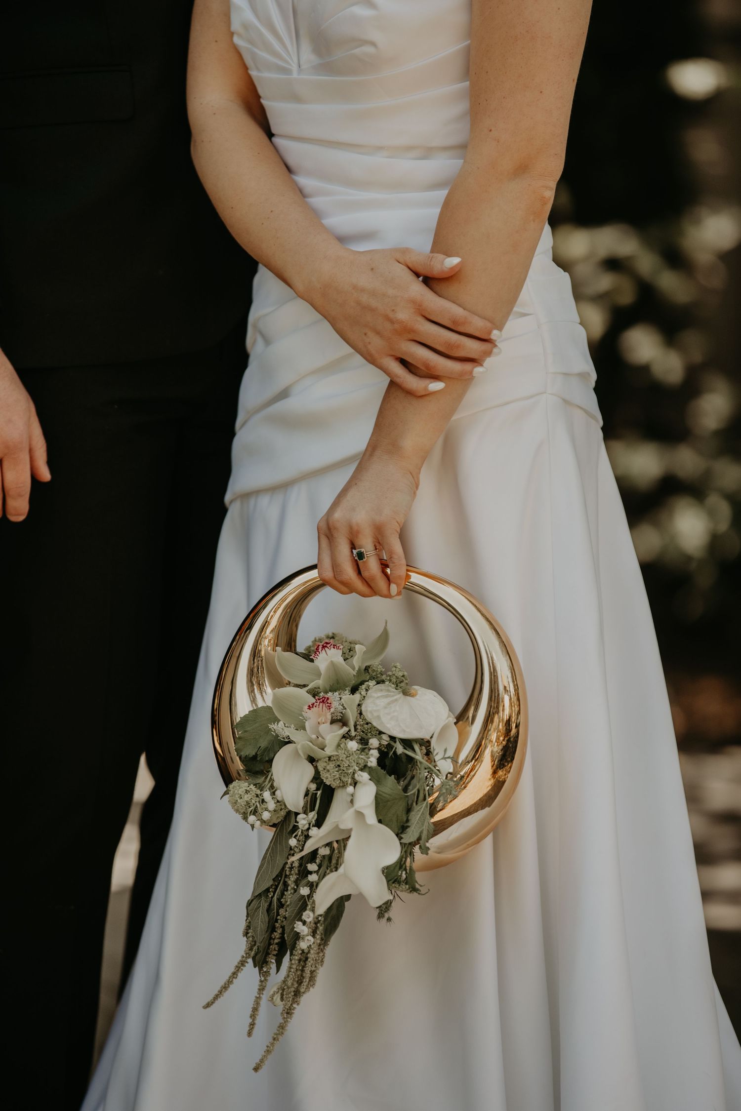 A person in a white wedding dress holds a modern metallic gold circular bouquet holder with white flowers and greenery.