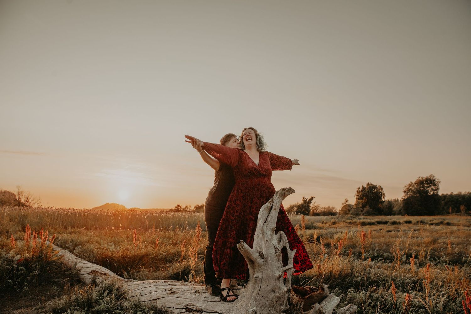 A figure in a flowing red dress stands with arms outstretched on a fallen tree at sunset in a rural landscape.