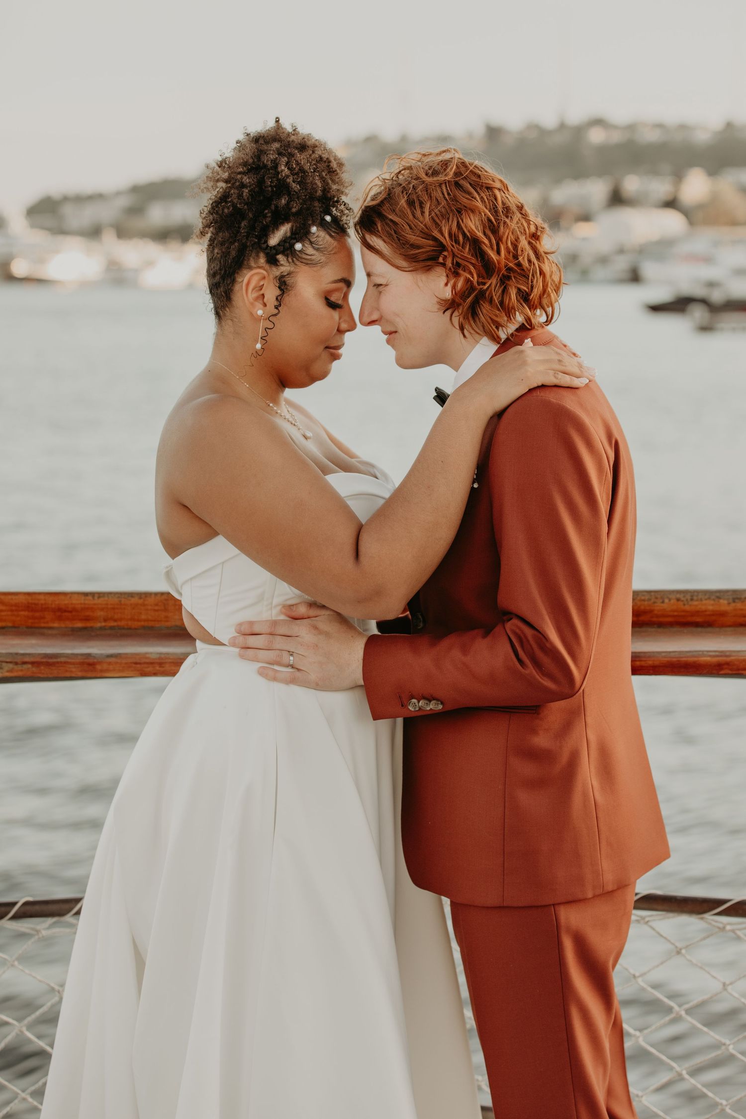 A romantic couple embraces on a boat dock with water and boats visible in the background.