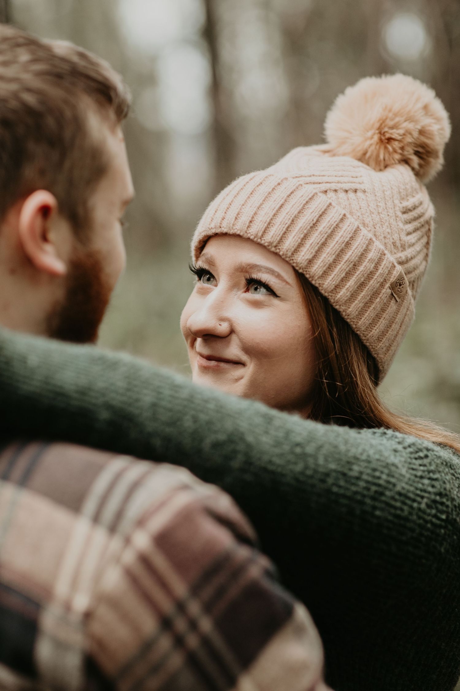 A person wearing a beige knit beanie with pom pom smiles warmly during an autumn outdoor photoshoot.