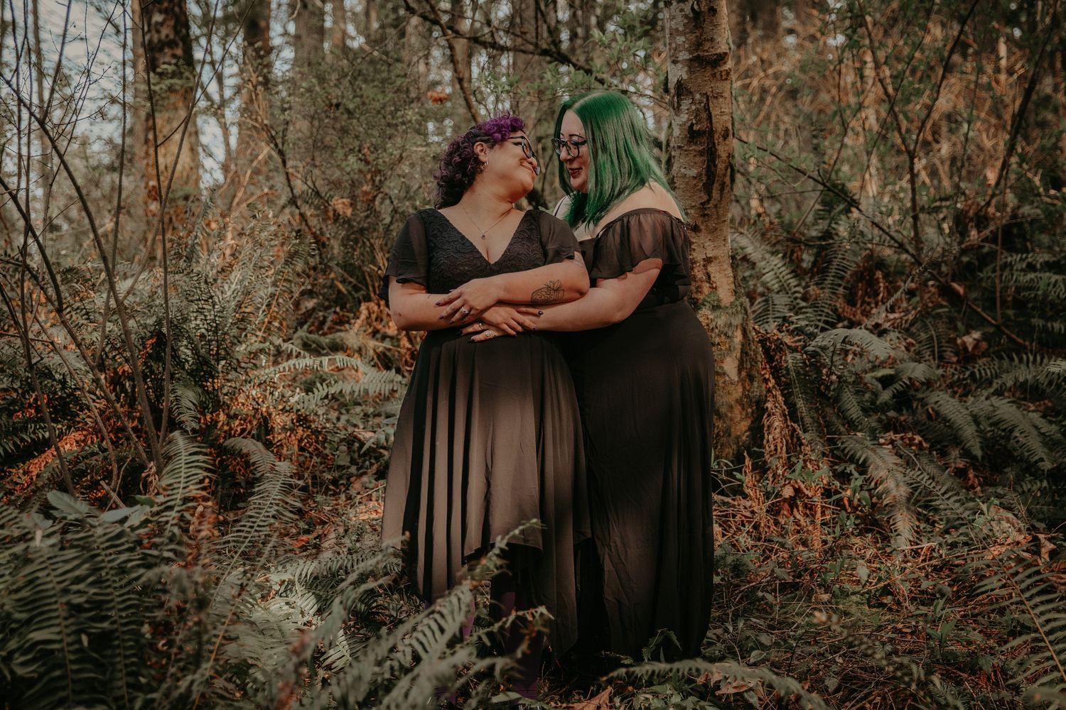 Two people in dark dresses embrace intimately in a moody forest setting with ferns and fallen leaves.