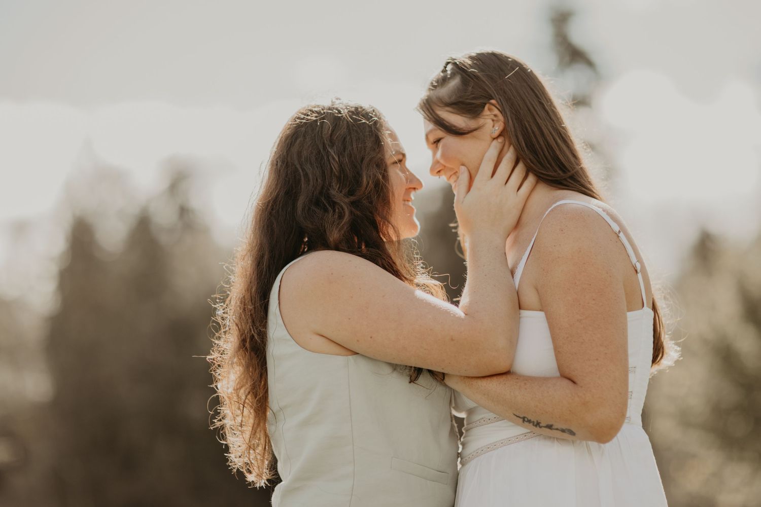 Two people in light-colored clothing share an intimate moment at sunset with trees in background.
