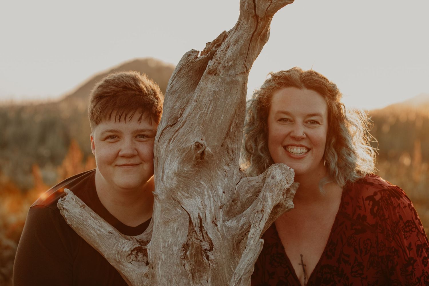 Happy couple posing next to driftwood during golden hour photoshoot.