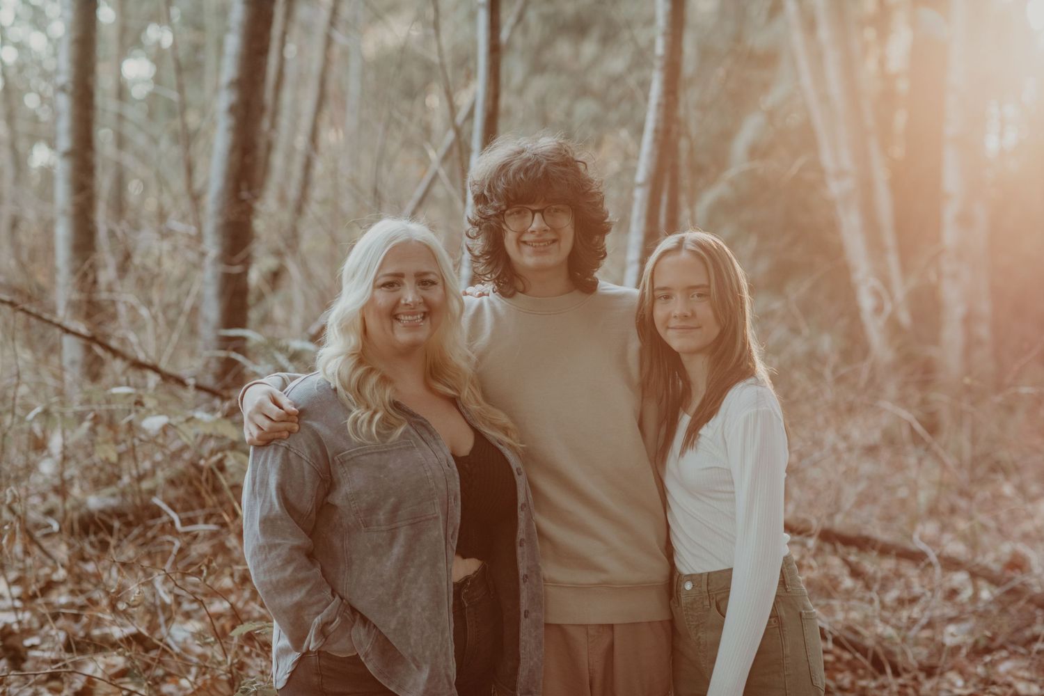 Three friends stand close together in a sunlit wooded forest area wearing casual autumn clothing.
