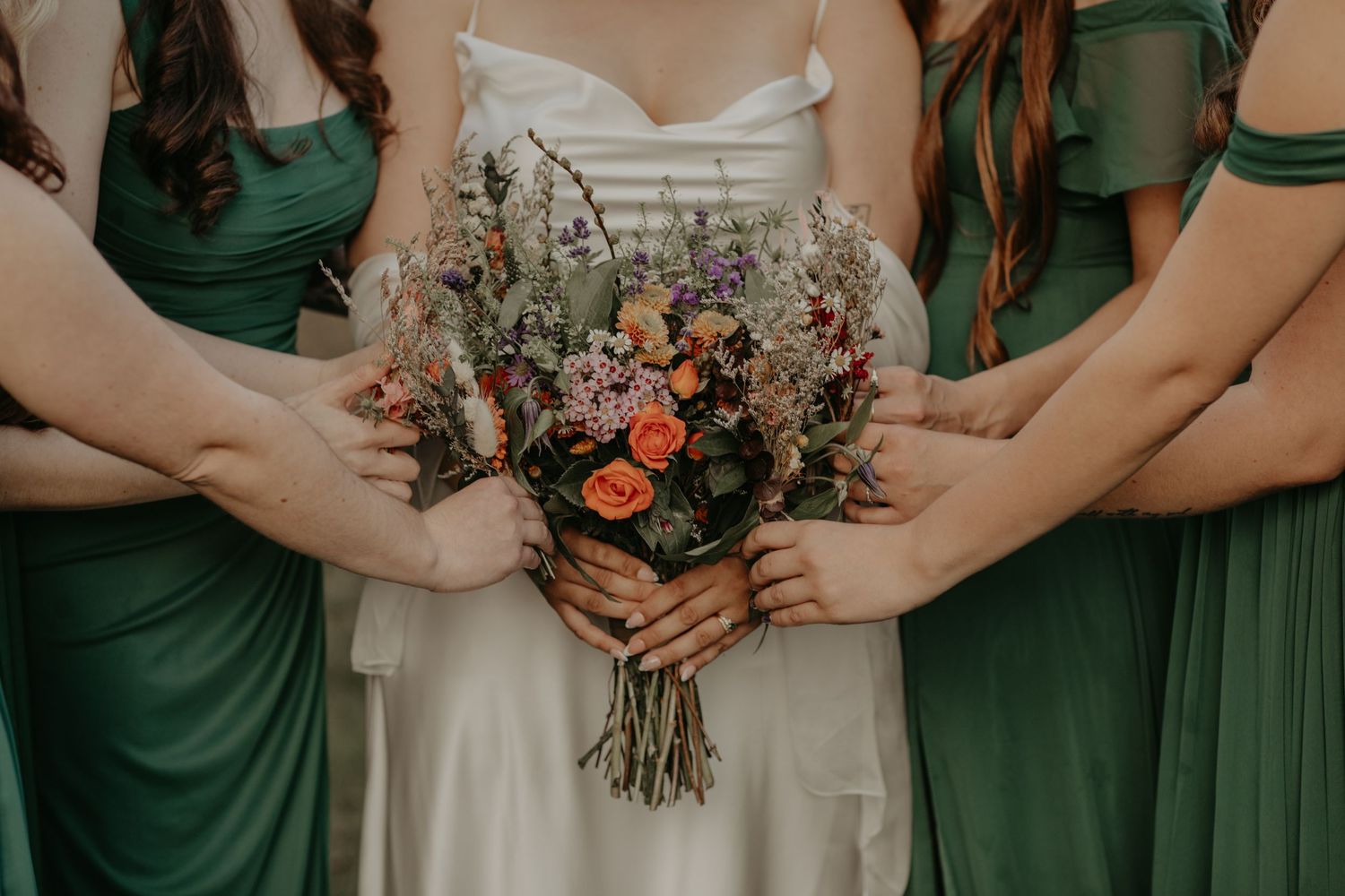 Wedding bouquet featuring coral roses and wildflowers held between people wearing emerald green and white dresses.