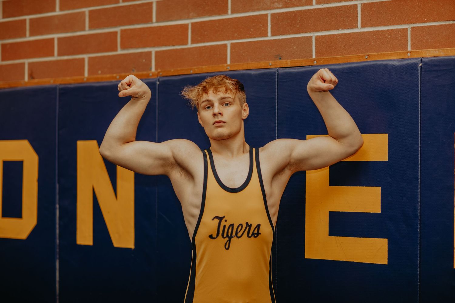 A wrestler in a yellow singlet with 'Tigers' text flexes their arms against a blue and orange wall backdrop.