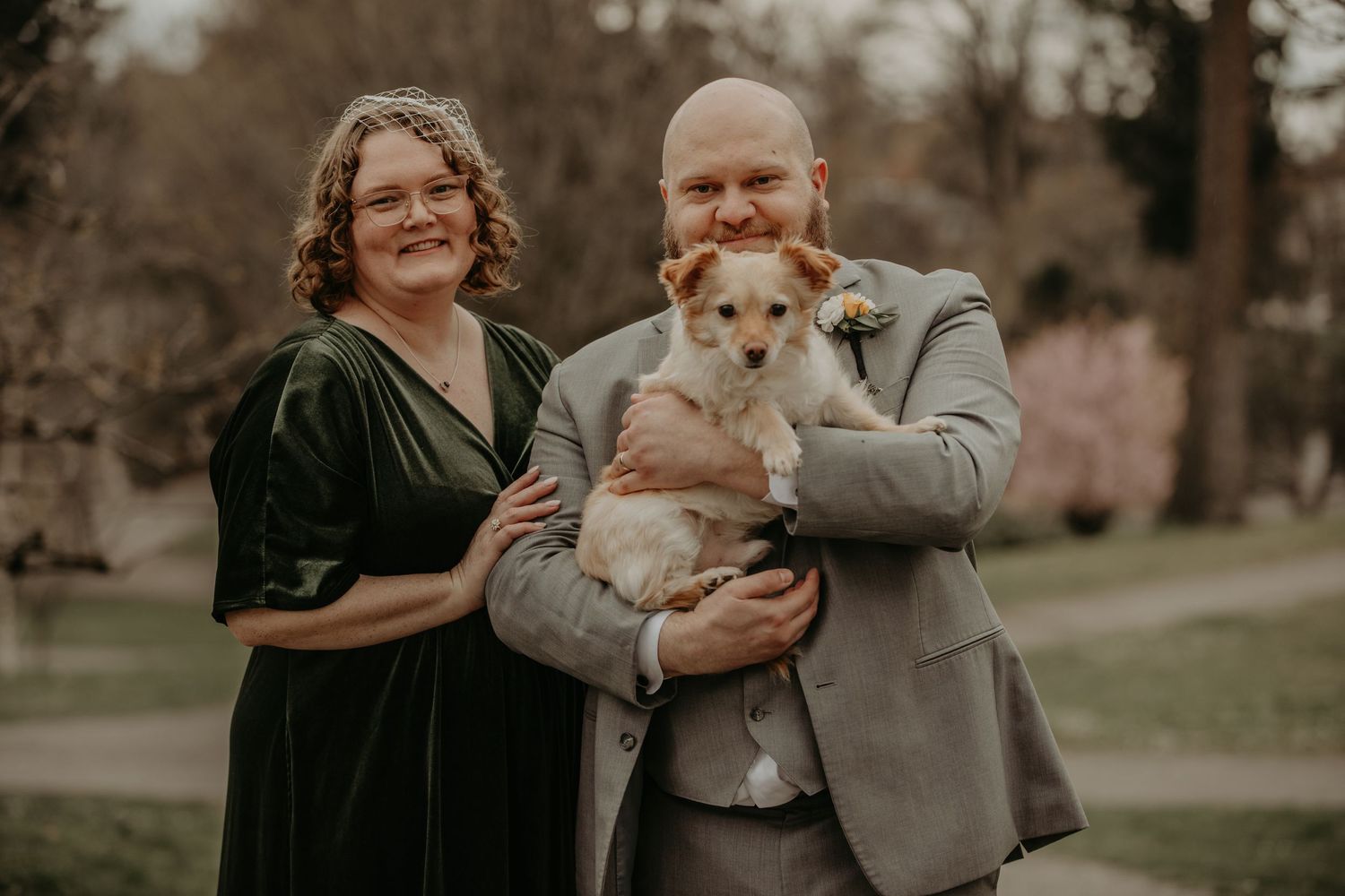 A couple stands outdoors in formal attire on a grassy lawn while holding their small brown and white dog.