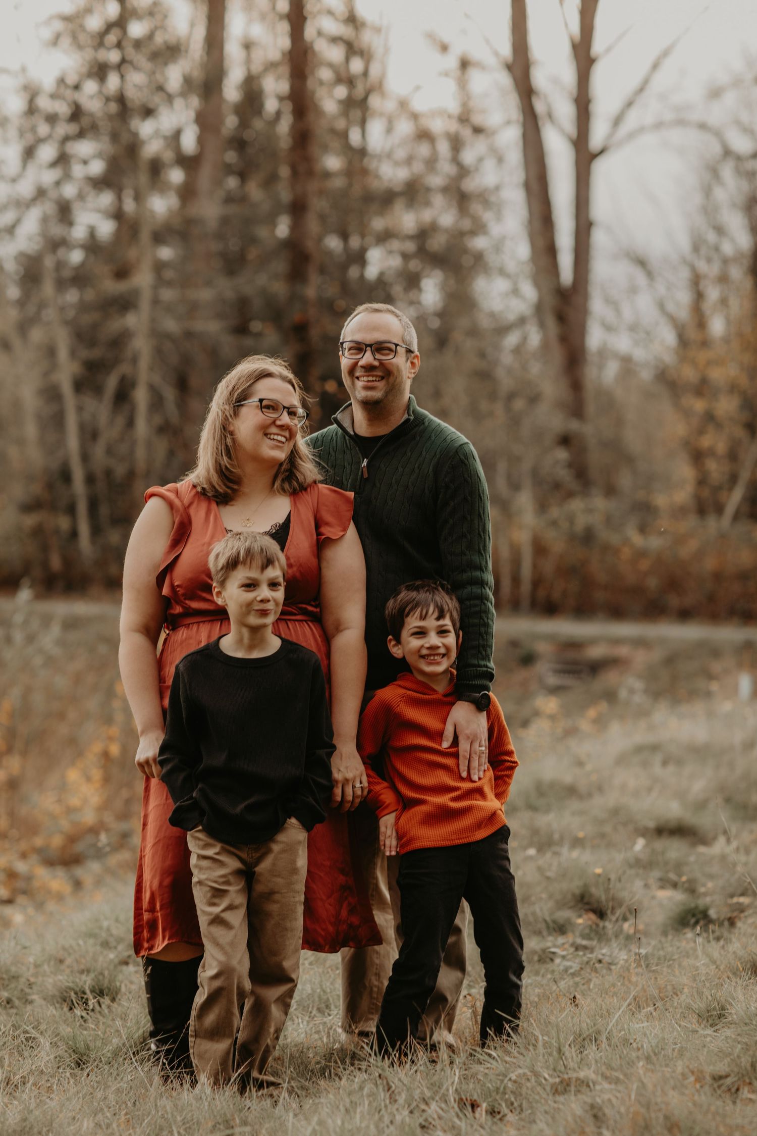 A family of four stands together outdoors in coordinating red and black outfits against an autumn forest backdrop.