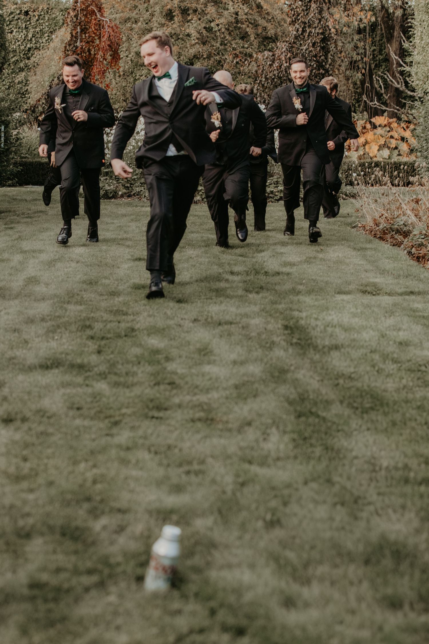 Groomsmen in dark suits run across a green lawn chasing after a bottle while having fun at a wedding event.