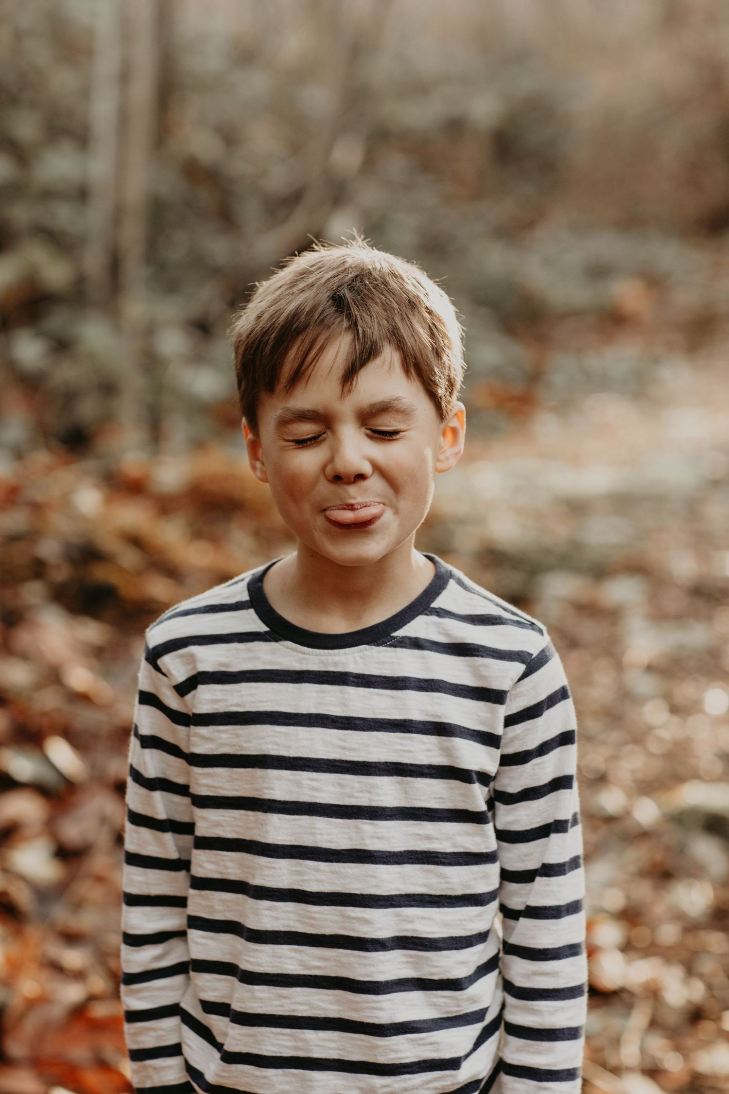 Child wearing a black and white striped long sleeve shirt smiles with eyes closed in an autumn forest setting.