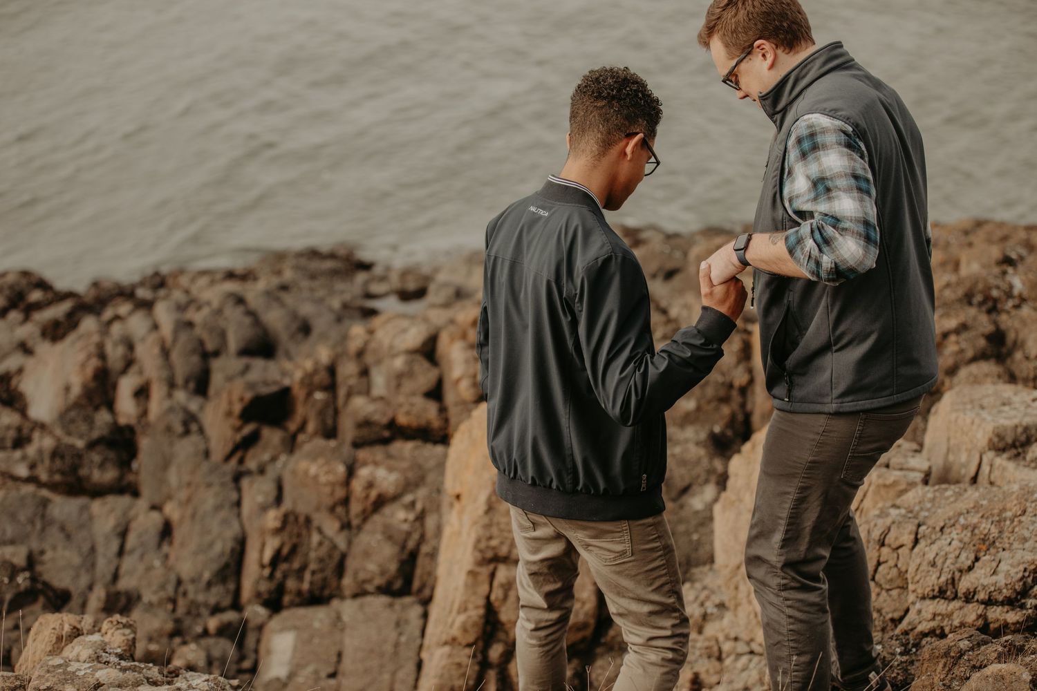 Two people in casual jackets stand on rocky terrain examining something together near a shoreline at dusk.