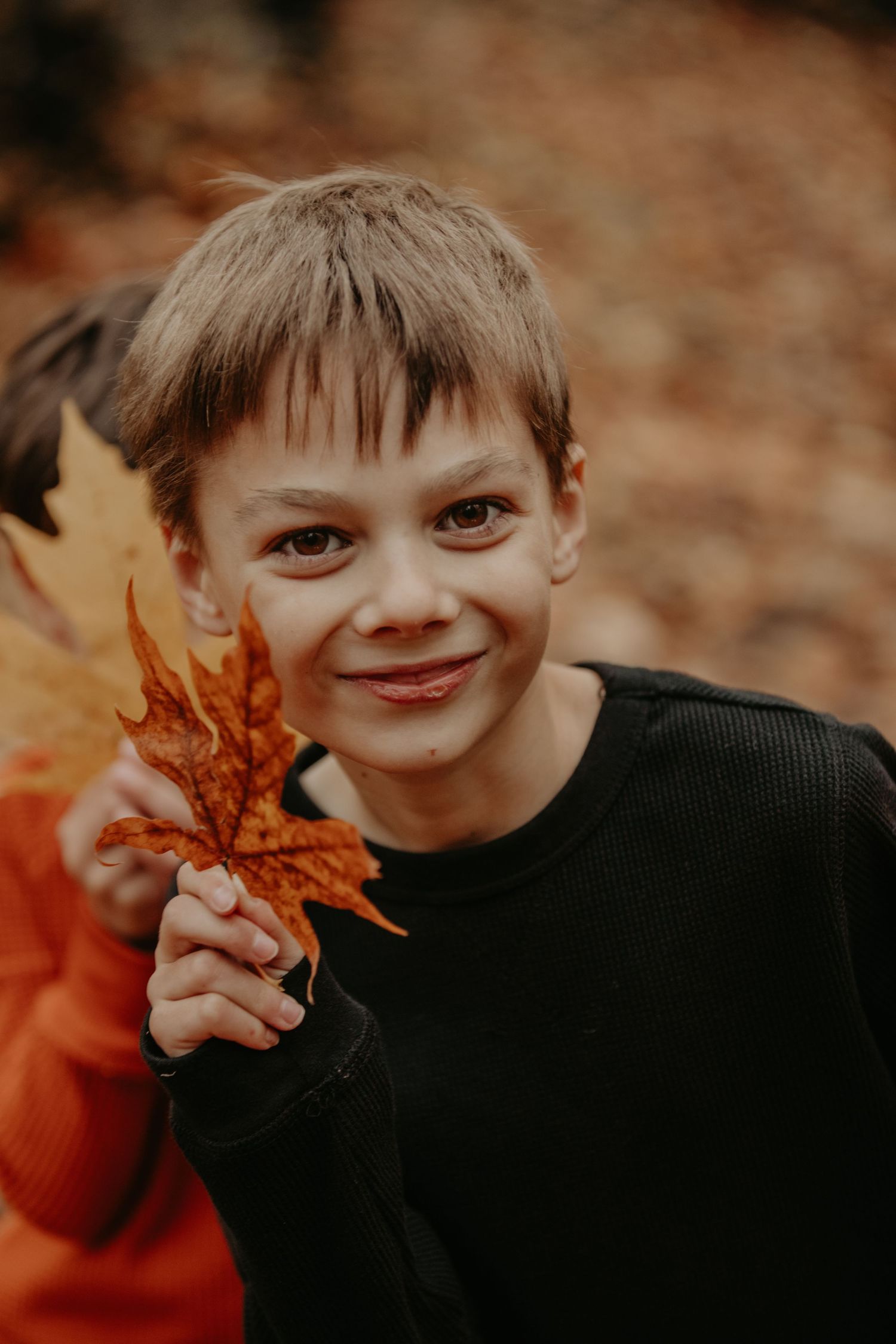 A smiling child in a black sweater holds up a bright orange autumn leaf outdoors.