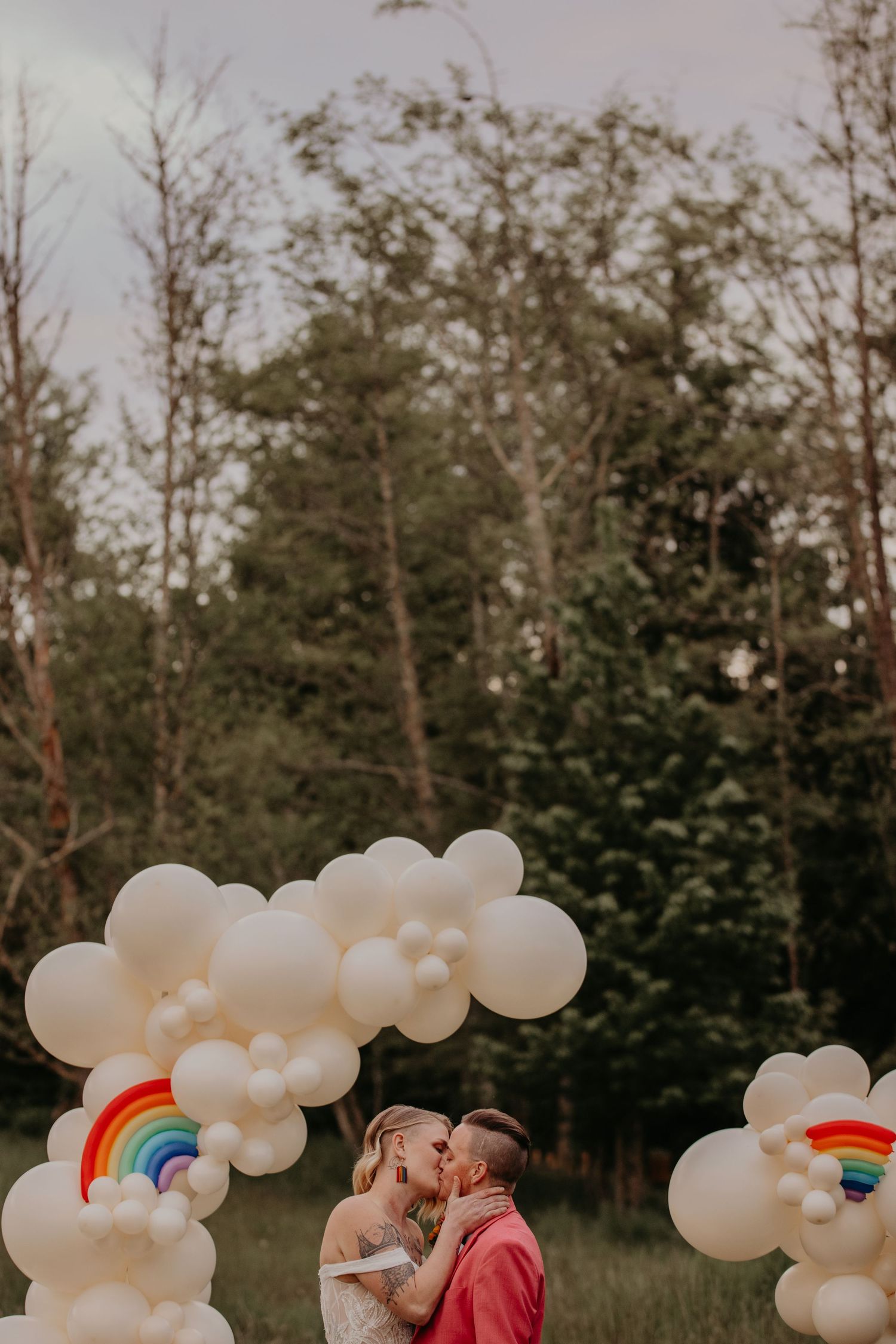White balloon arch with rainbow accents decorates an outdoor photoshoot in a wooded setting.