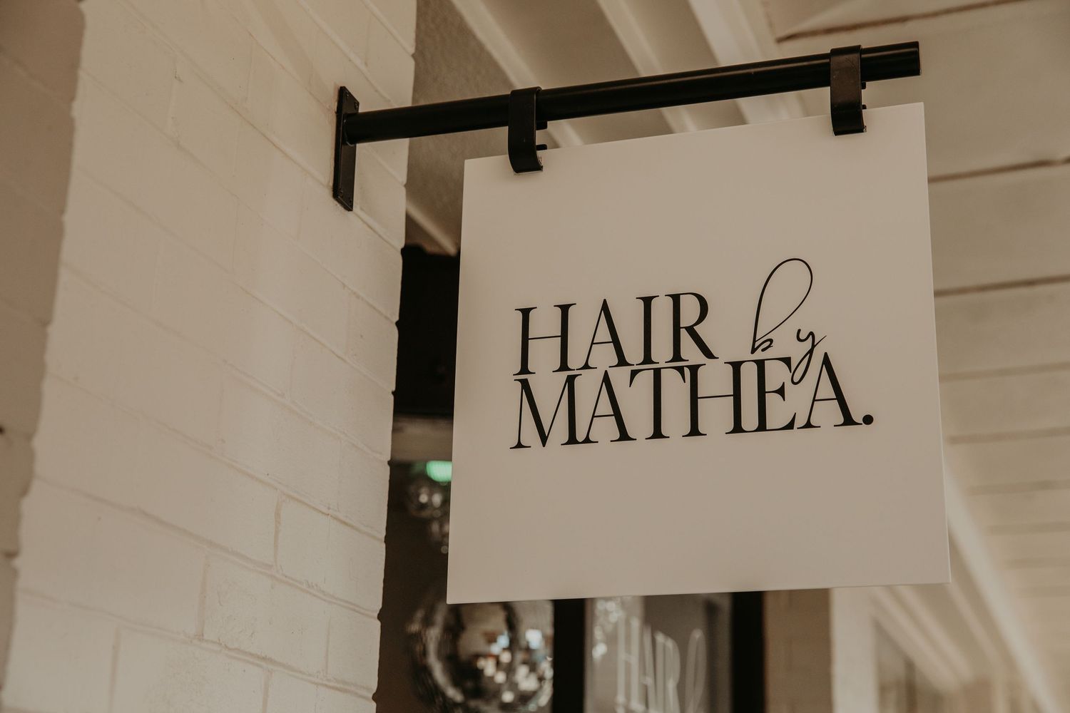 A black and white hanging business sign for Hair by Mathea against a white exterior wall.