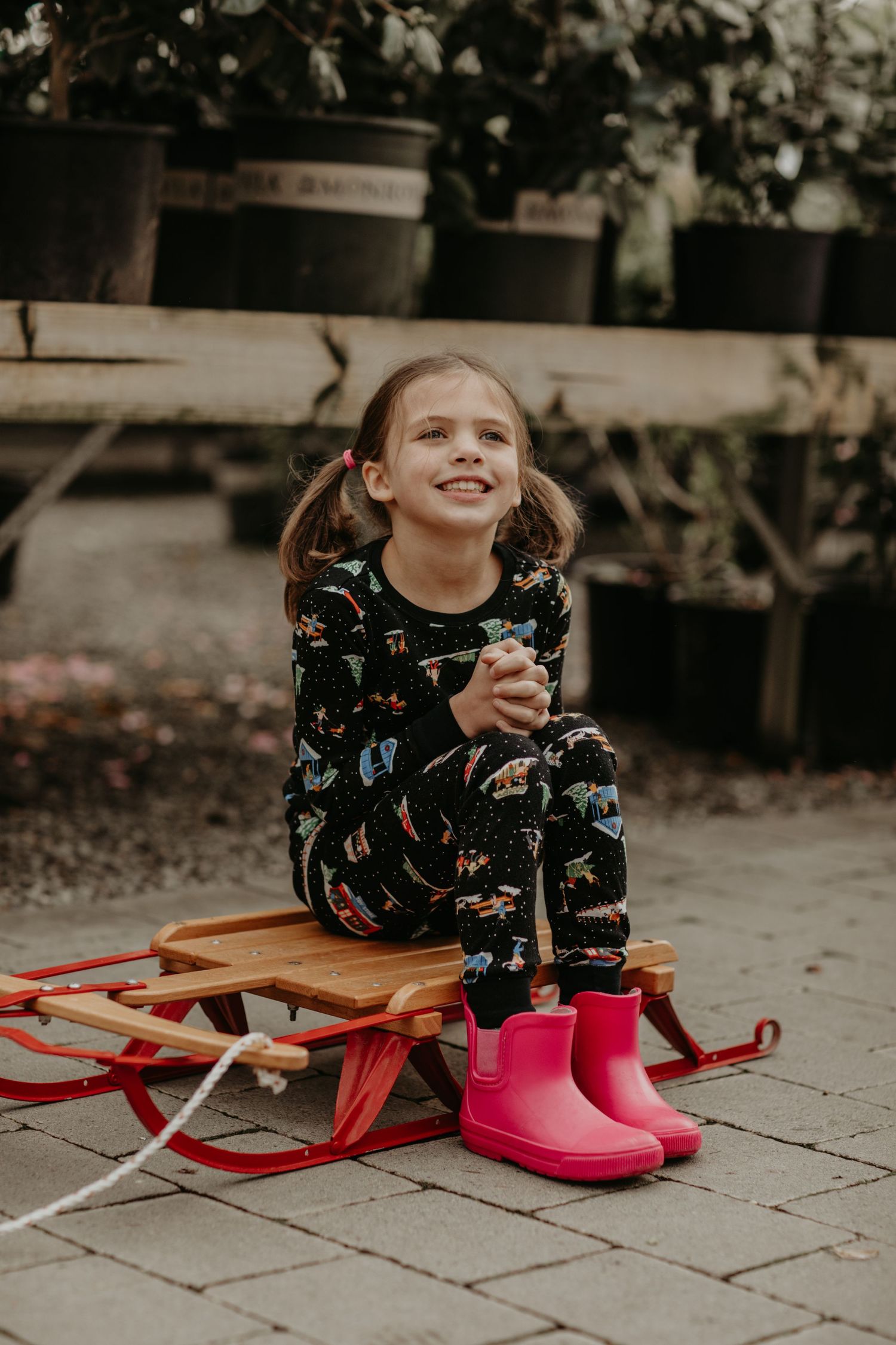 Child sits on vintage red wooden sled wearing dinosaur pajamas and bright pink rain boots in outdoor garden setting.